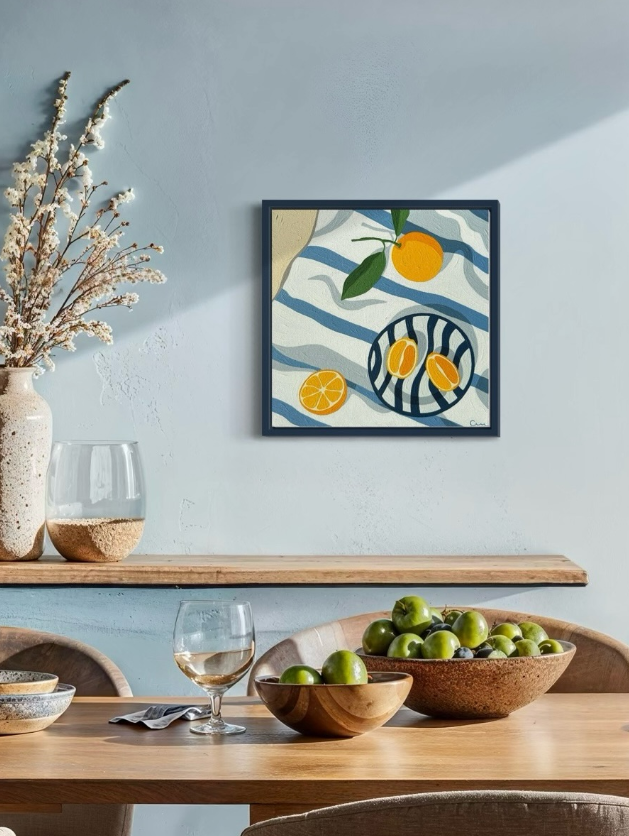A dining room with a wooden table, a bowl of green apples, a glass of white wine, and a glass container of water with a plant beside it. A colorful still life artwork of citrus fruits and a striped pattern hangs on the light blue wall.