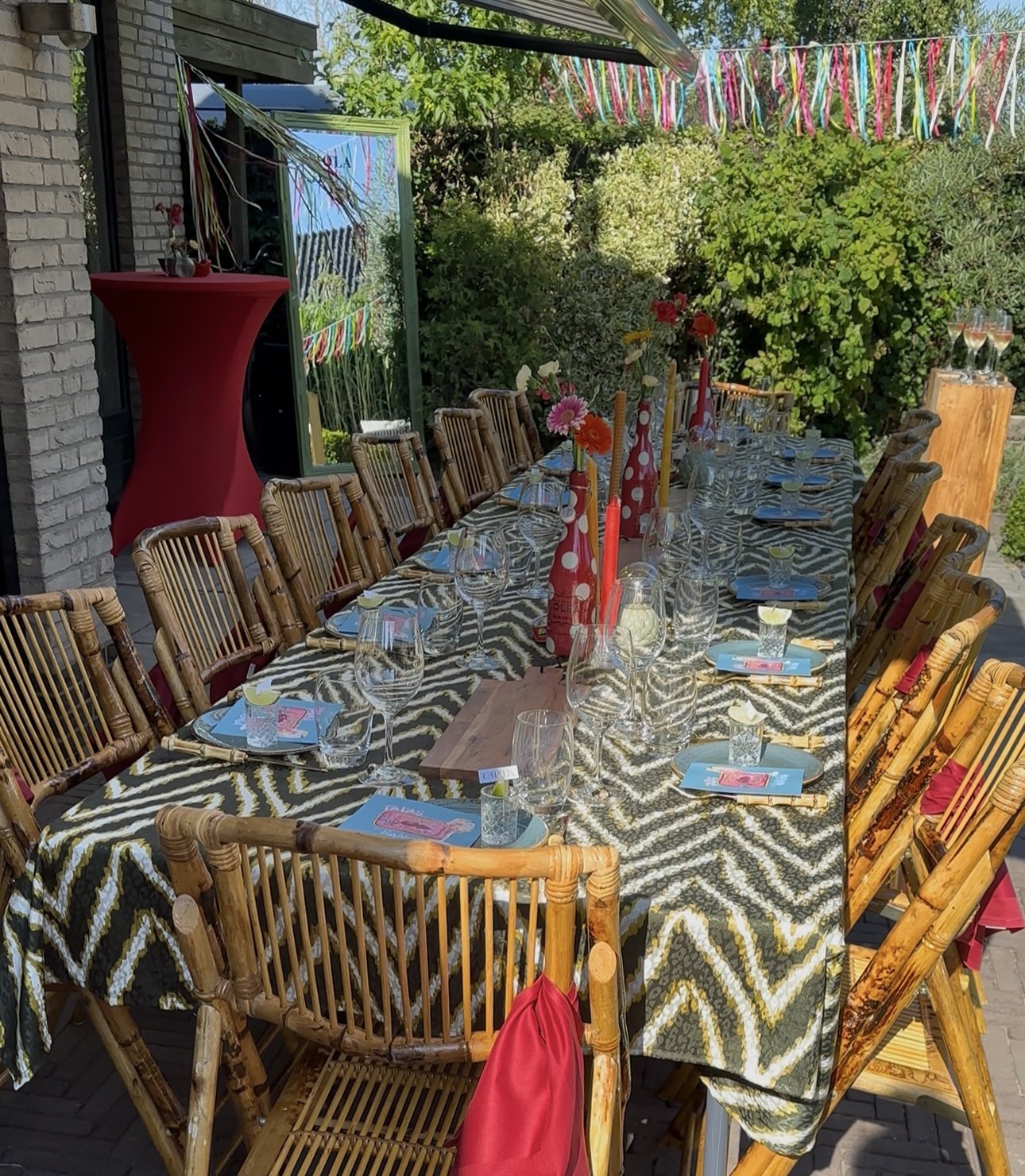 Bruidstafel met decoratie, inclusief gele en rode slingers, bloemen, en drankjes, buiten onder een blauwe lucht met groene struiken op de achtergrond.