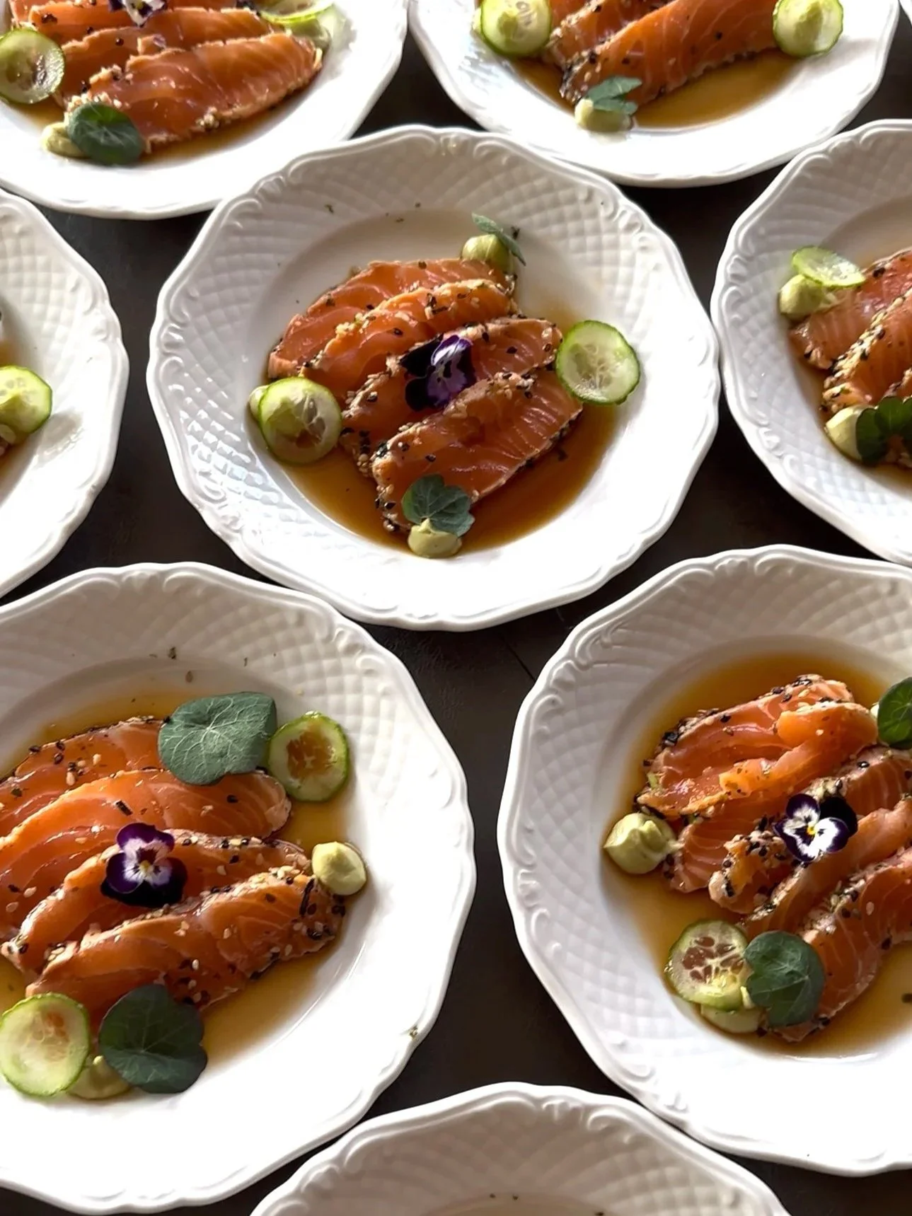 Fijn gesneden rauwe zalm op witte borden, gegarneerd met groene komkommers, paarse viooltjes, kleine groene bladeren, sesamzaadjes en een lichtbruine saus.