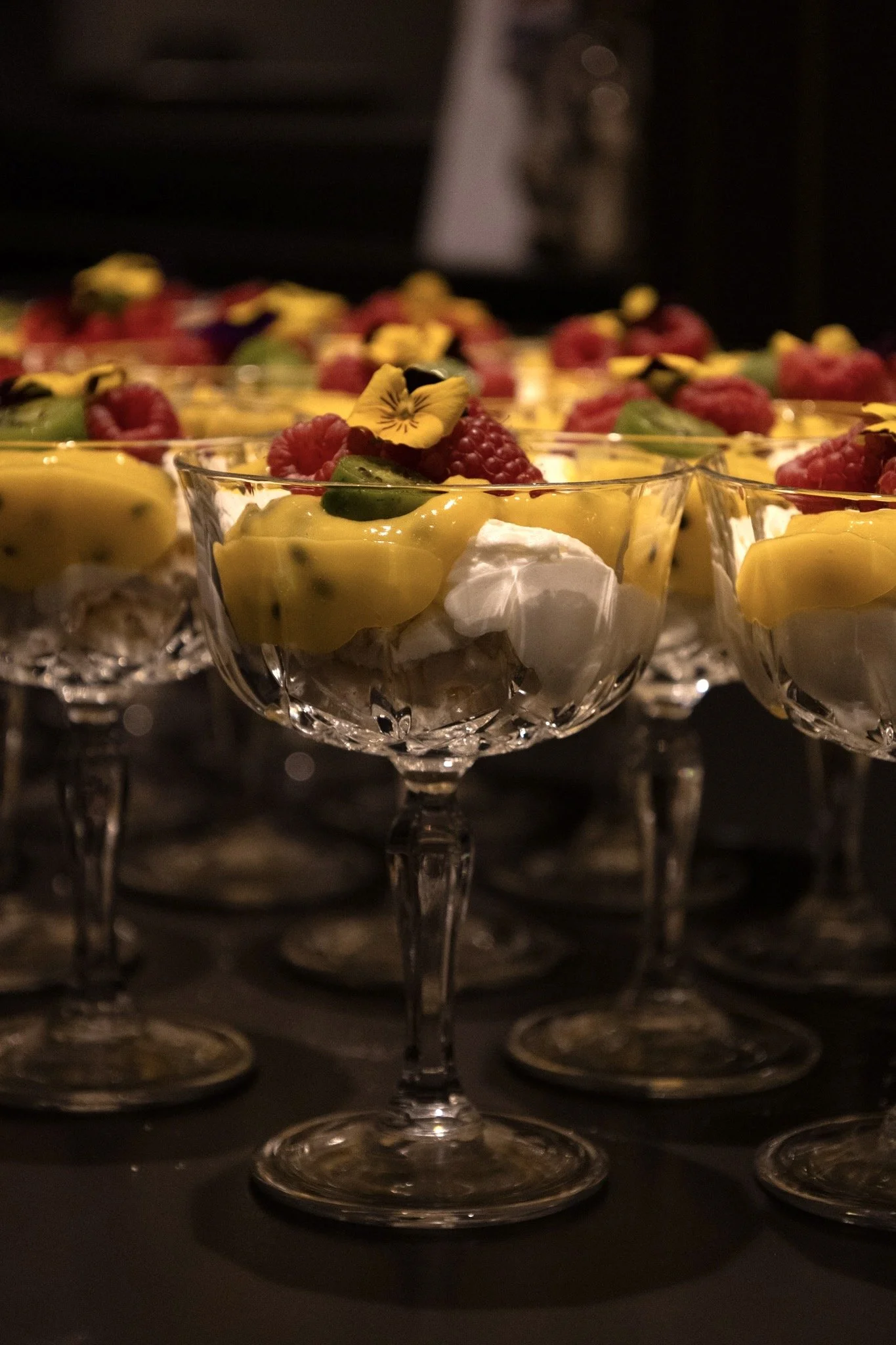 Fotografie van meerdere glazen dessertkommen gevuld met smaakvolle desserts, versierd met fruit en eetbare bloemen, uitgestald op een donkere achtergrond.