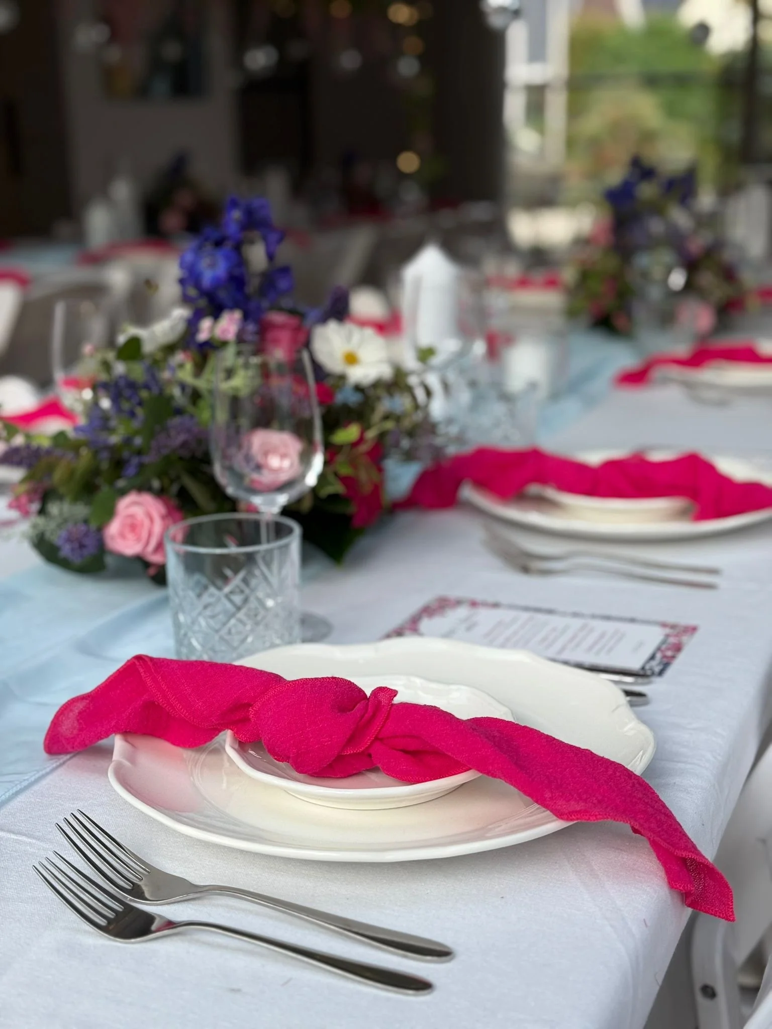 Een tafel met bestek, een witte bord met een roze servet eromheen, een vaas met bloemen, glazen en menu's op een wit tafelkleed.