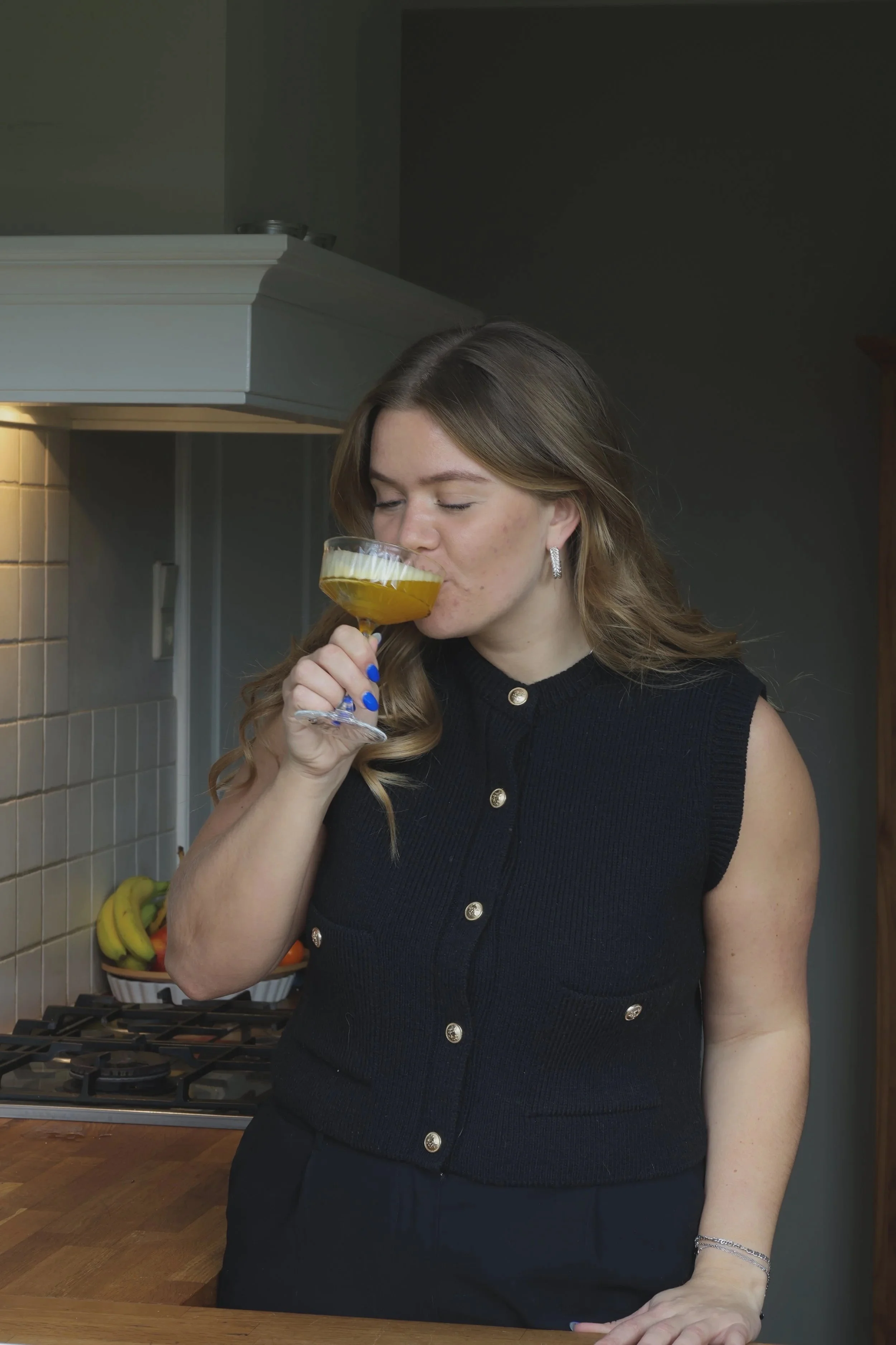 Een vrouw drinkt een glas thee in een keuken, met een basket met bananen en andere fruit op de achtergrond.
