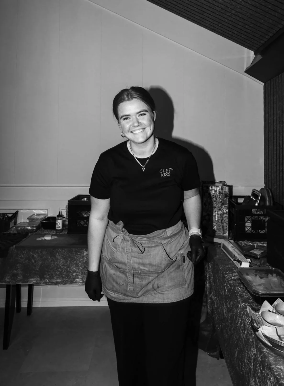 Een vrouw in een zwart T-shirt en een schort staat achter een tafel in een keuken of cateringruimte, lachend naar de camera.