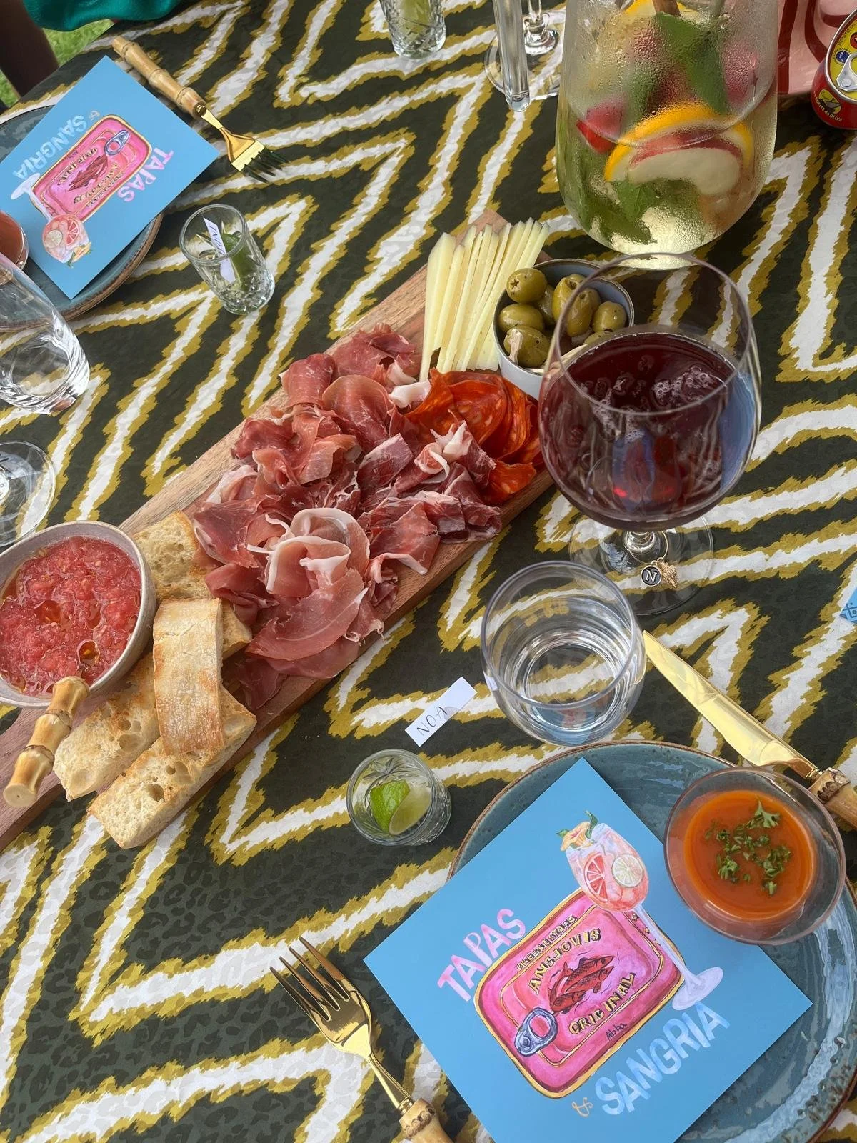 Een dienblad met verschillende koude vleeswaren en kaas, omringd door drankjes en snacks op een tafel met een zebraprint tafelkleed.