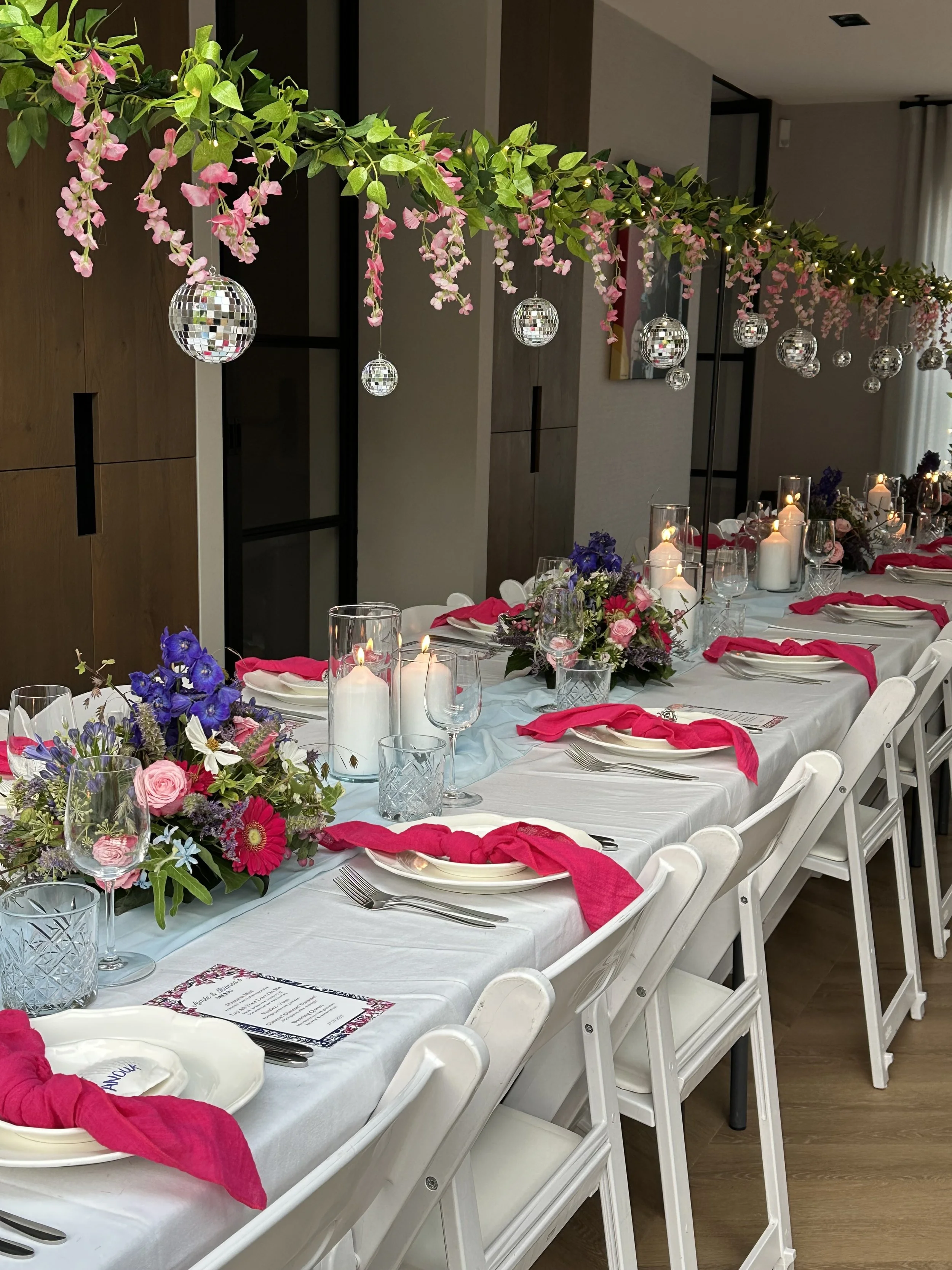 Een feesttafeldecoratie met een witte tafel, bloemstukken in glazen vazen, gedekte borden, bestek, glazen en rode servetten. Hangend boven de tafel hangen decoratieve ballen en groene takken met roze bloemen, met lampjes er tussen.