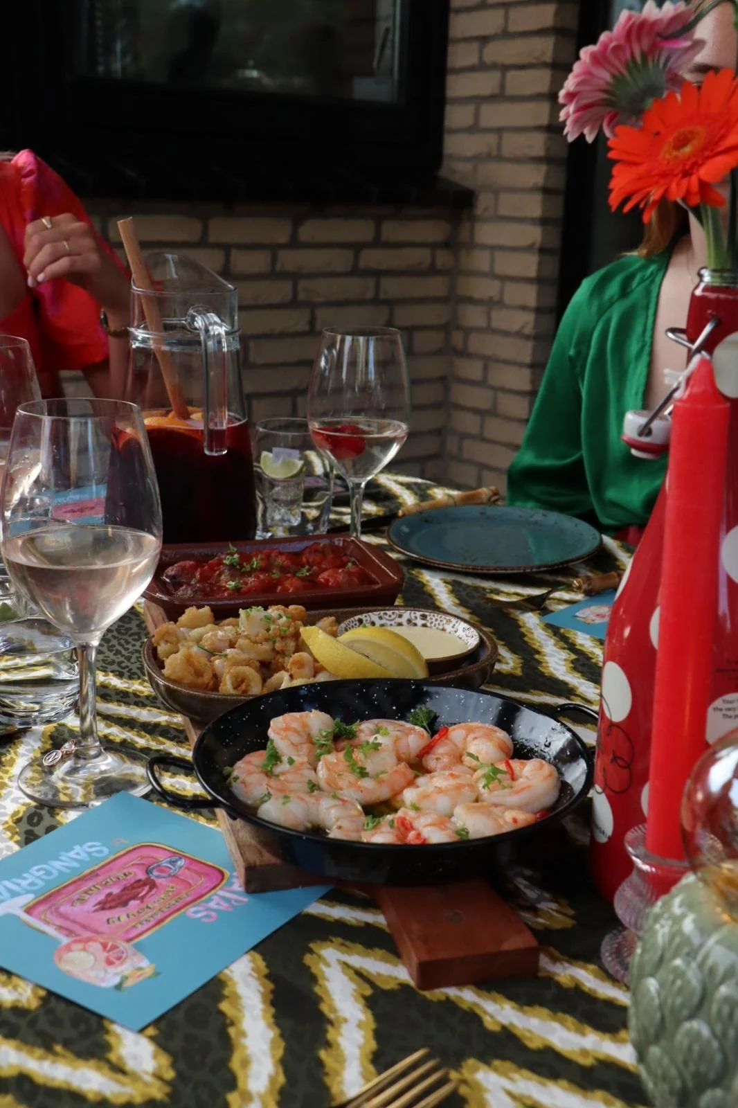 Een tafel met verschillende etensgerechten en glazen water en wijn, een fles met rode saus, bloemen in een vaas, en mensen aan de tafel, met een bakje garnalen, gekookte calamari, een glas met een roze drankje, en een glas met water, op de achtergrond een bak met tomatensaus.