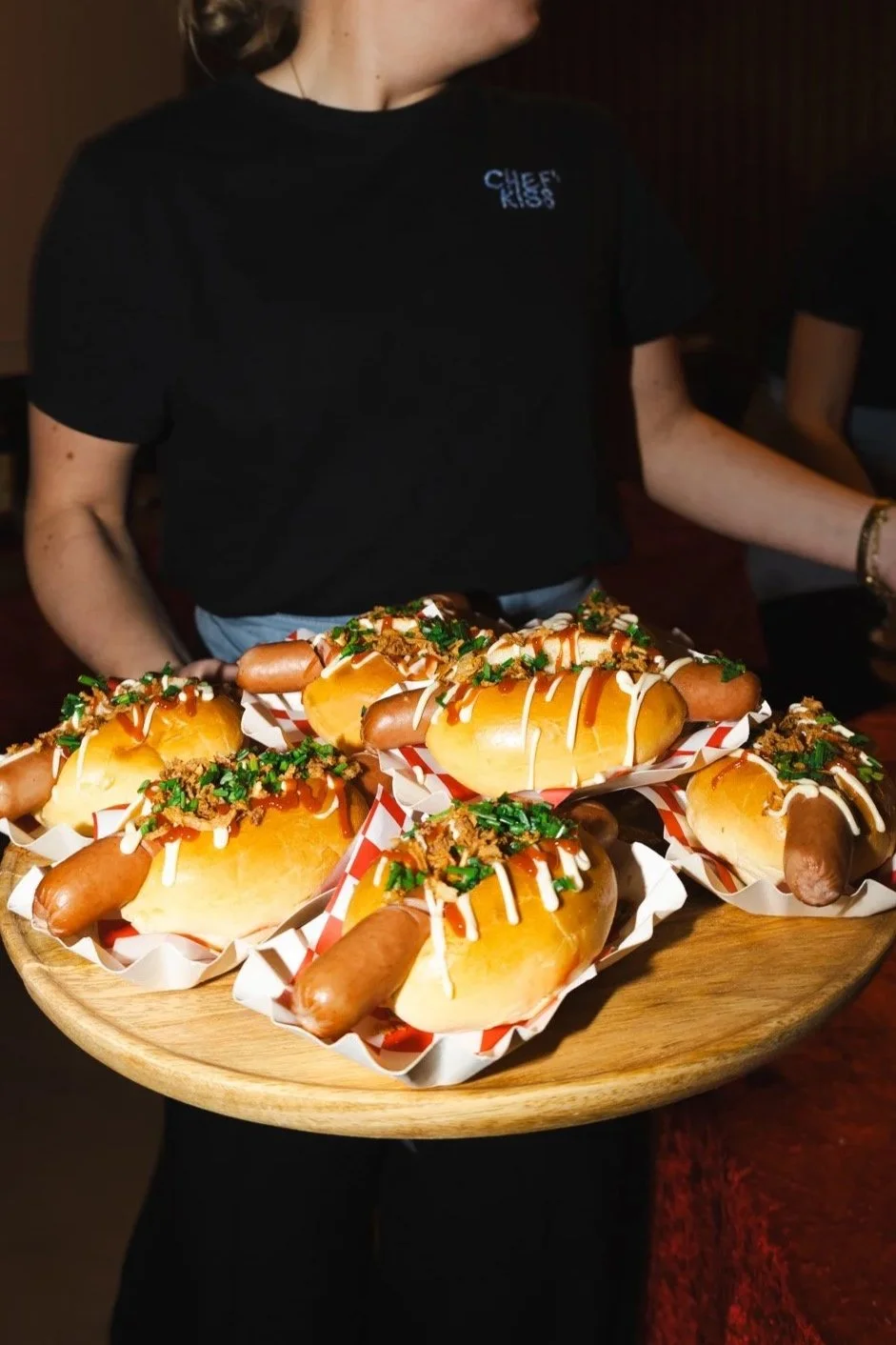 Persoon houdt een grote ronde houten serveerschaal met hotdogs met broodjes, worst, saus, gesneden groene kruiden en toppings.