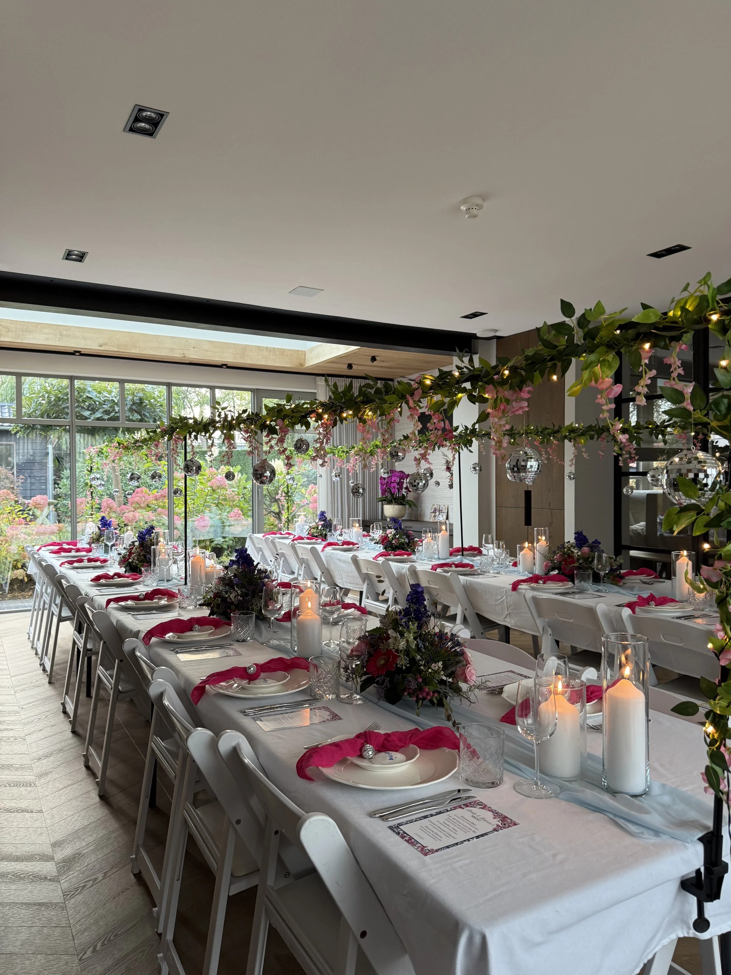 Een feestzaal ingericht voor een diner met lange tafels, witte tafellakens en roze servetten. Decoraties met bloemen en kaarsen, en hangende disco-ballen door de ruimte. Grote ramen met uitzicht op een tuin en bloemen buiten.