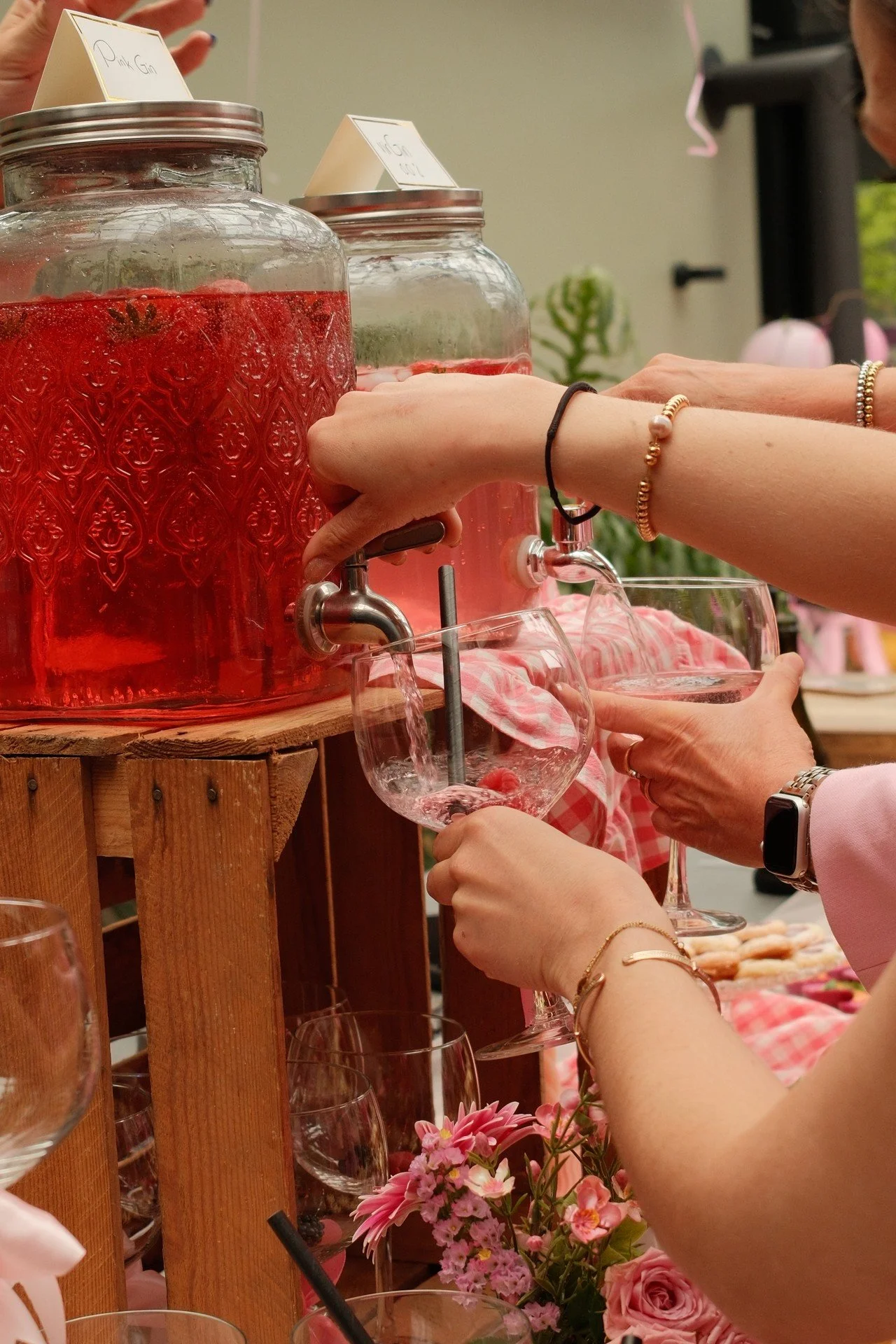 Mensen serveren roze limonade uit grote glazen waterkannen op een feestje met bloemen en decoraties.