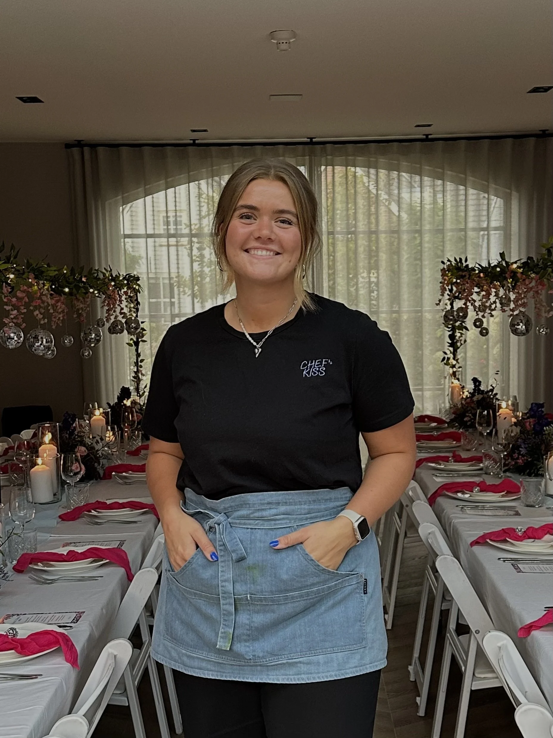 Een jonge vrouw in een zwarte T-shirt met de tekst 'Chef's Kiss' en een denim rok staat in een feestzaal met versierde tafels en verlichting.
