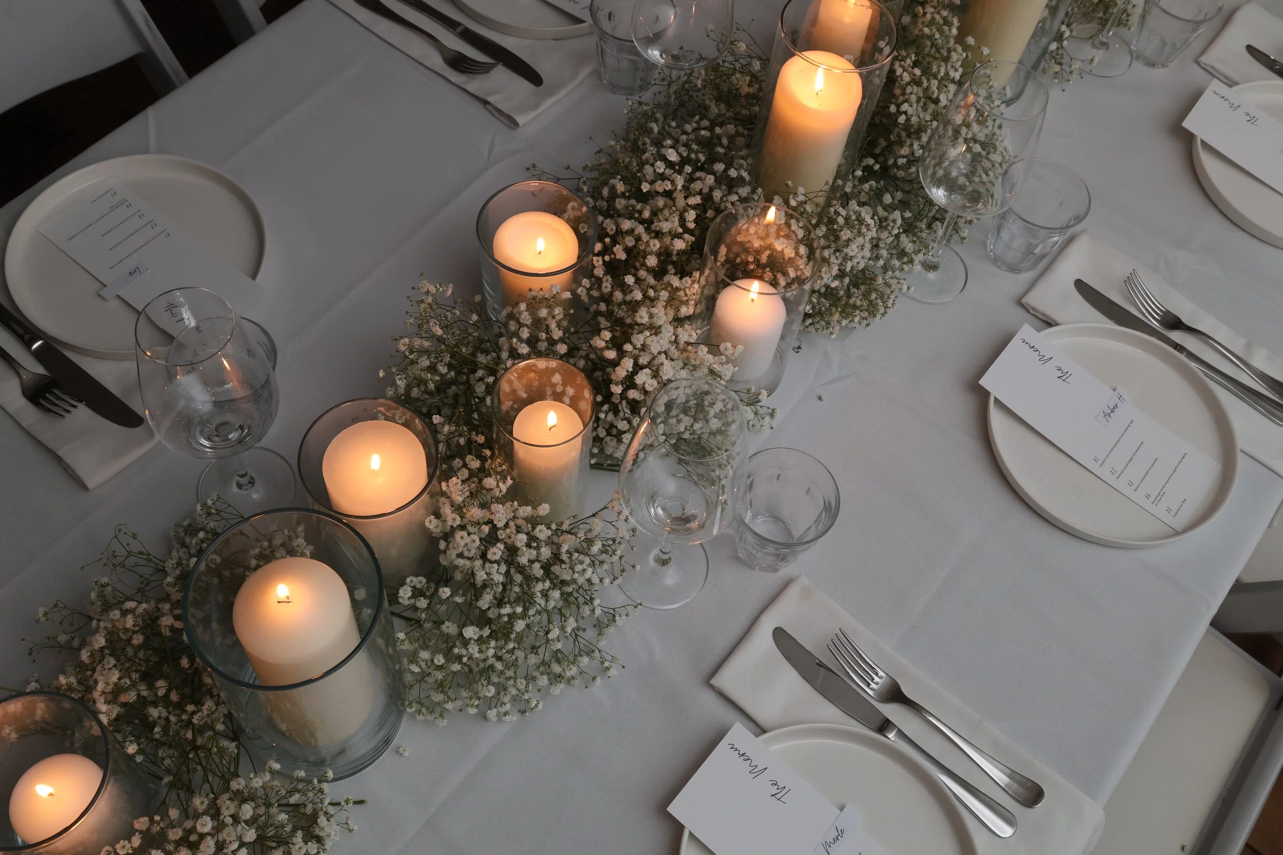 Een elegante dinertafel versierd met witte tafellaken, kaarsen in glazen houders, kleine witte bloemen en tafelinstellingen met wit bord, mes, vork, lepel, en menu-kaarten.