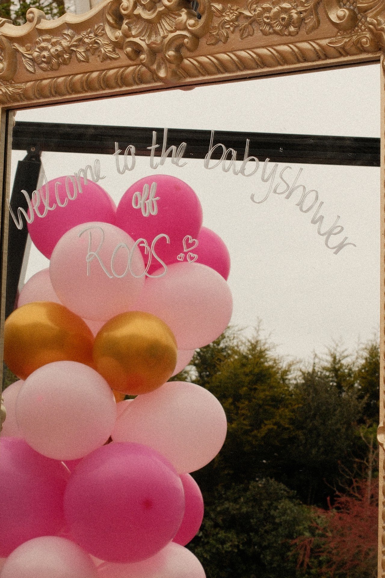 Een decoratief bord met een spiegel die reflecteert, met daarop een tekst en een cluster van roze, gouden en witte ballonnen. Op de spiegel staat geschreven: "Welcome to the babyshower of ROOS".