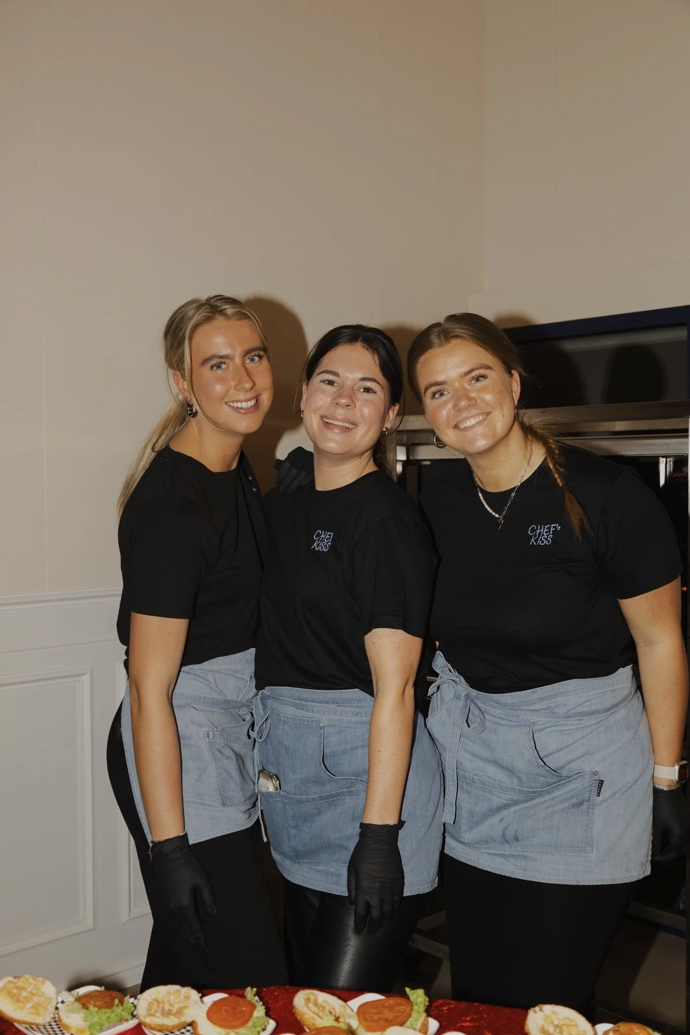 Drie vrouwelijke personen in zwart T-shirts en blauwe schorten die zich voorbereiden voor het serveren van etenskruiden en sandwiches.