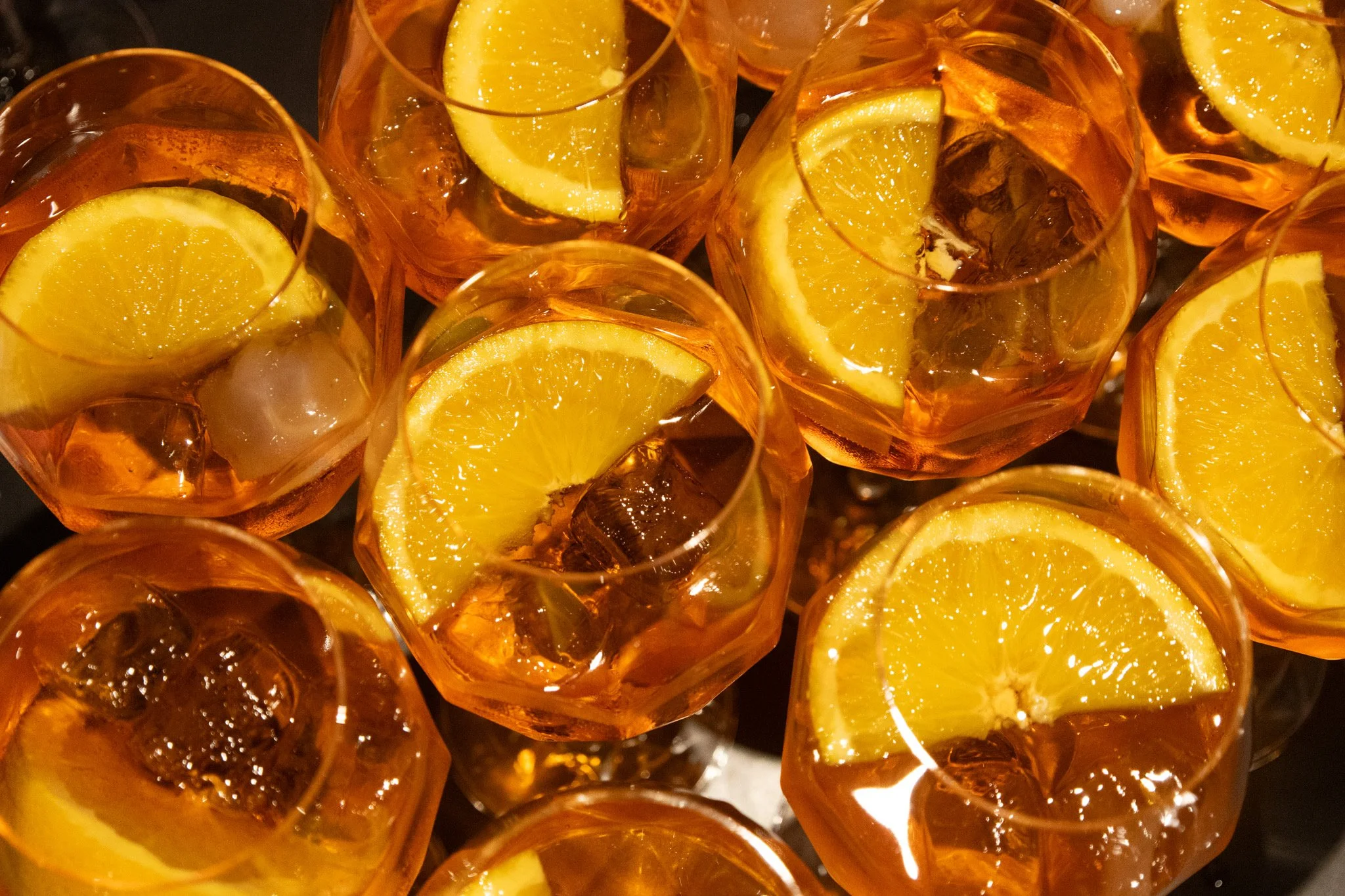 A group of glasses filled with an orange-colored drink, each garnished with a lemon wedge and ice cubes.