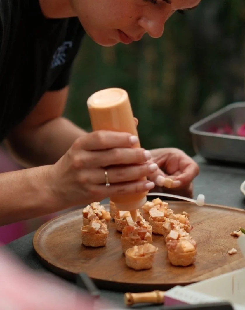 Persoon bereidt sushi met saus op een houten bord, andere ingrediënten liggen op de tafel