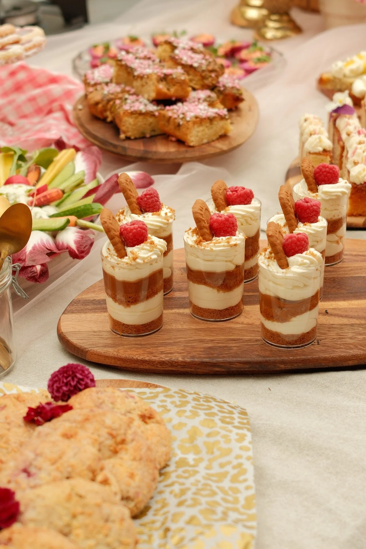 Miniatuur dessertglas met lagen van koek, room, en framboos, versierd met een koekje en een framboos op de top, uitgestald op een houten serveerplank.