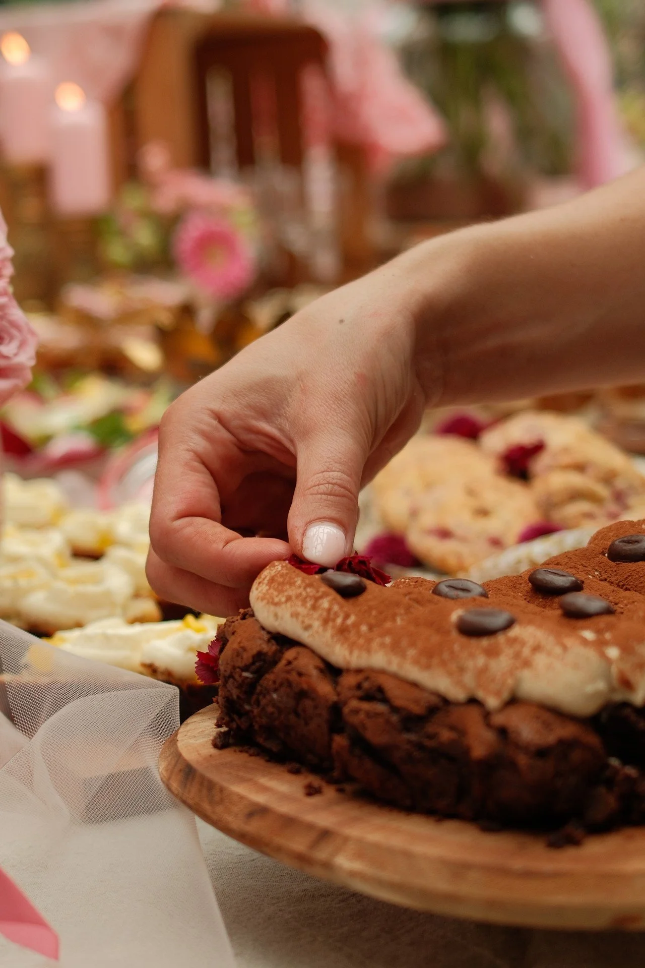 Hand die een stuk chocoladecake met rode garnering en chocoladestukjes optilt op een houten bord, met een versierd desserts buffet op de achtergrond.