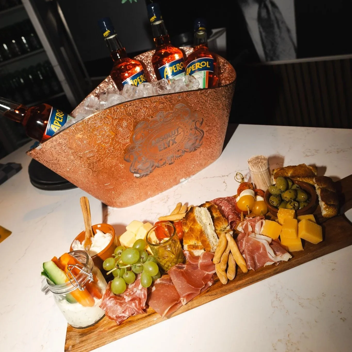 Een plaat met diverse hapjes zoals olijven, kaas, vleeswaren, brood, groenten en dips, en een emmer met ijskoude Aperol-biertjes.