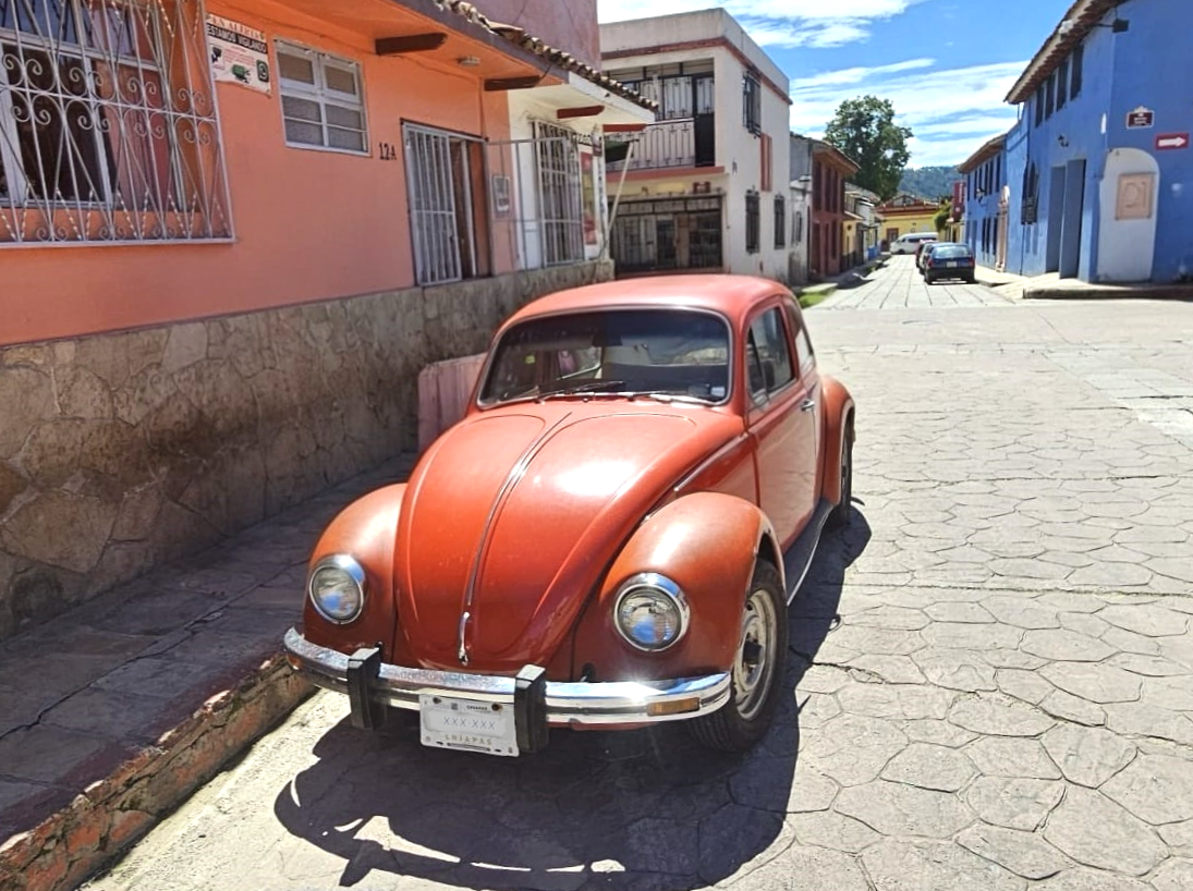 A vintage red Volkswagen Beetle car parked on a cobblestone street in a colorful neighborhood with pink, white, and blue buildings under a partly cloudy sky.