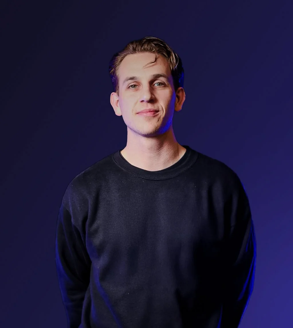 A young man with light skin and short, light brown hair, wearing a black sweatshirt, standing against a dark purple background with a slight smile.