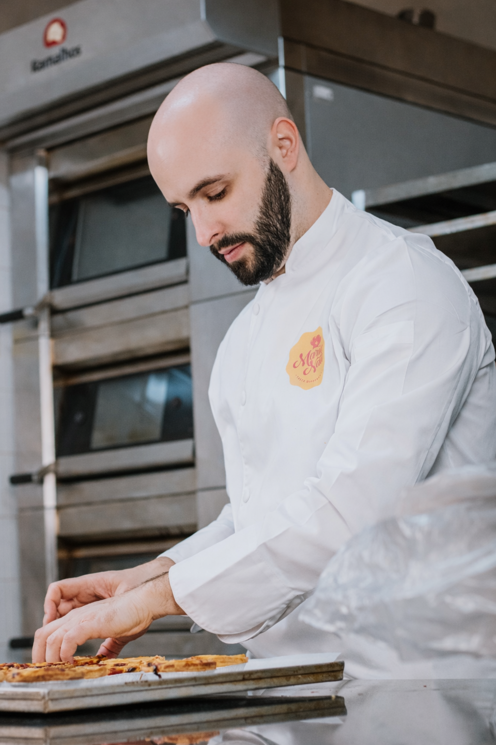 A chef with a beard, wearing a white chef's coat, is preparing pizzas in a professional kitchen.