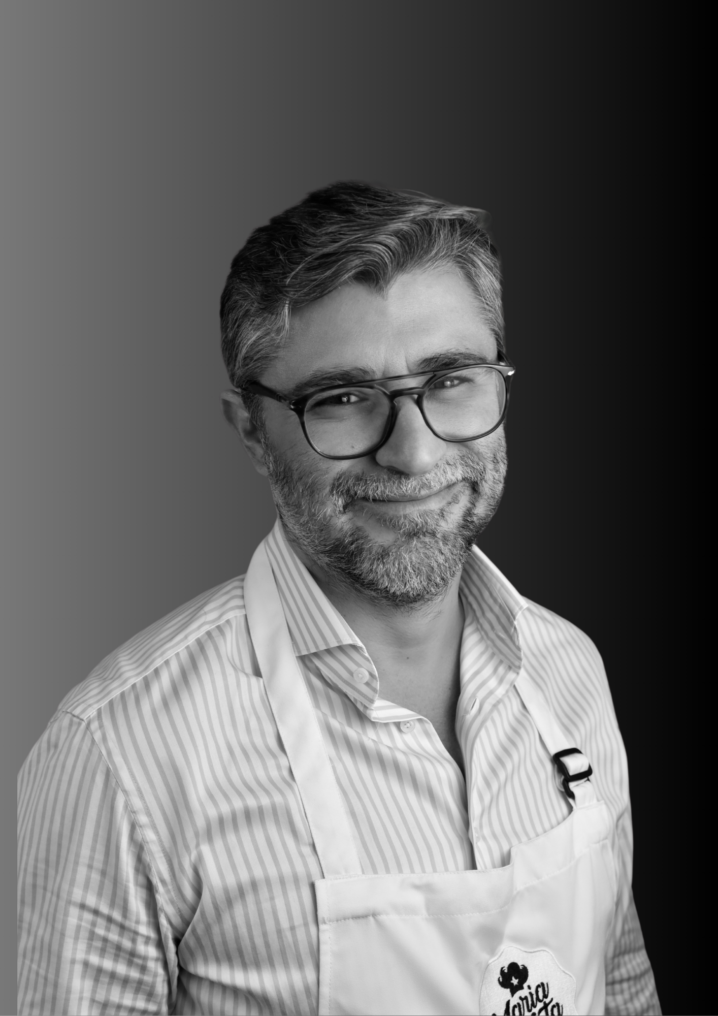 A black-and-white portrait of a smiling middle-aged man with glasses, gray hair, and a beard, wearing a striped shirt and a white apron with a logo.