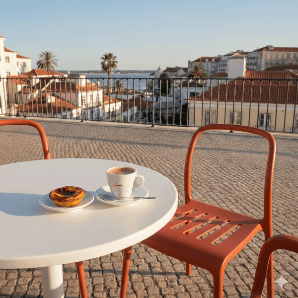 Outdoor terrace with a white round table, two orange chairs, a cup of coffee, and a cinnamon roll on a small plate, overlooking a scenic cityscape with rooftops and palm trees under clear blue sky.