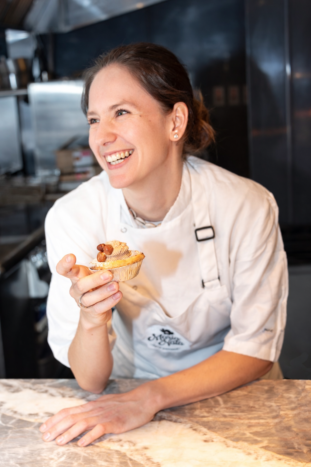 A smiling woman in a white chef's coat holding a cupcake with frosting and hazelnuts in a kitchen.