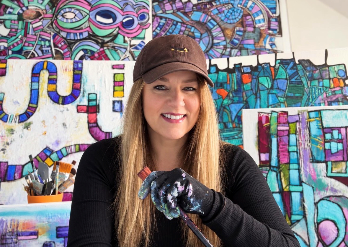 A woman with long blond hair wearing a brown cap and black long-sleeve shirt, smiling while holding a paintbrush in her hand, with colorful abstract paintings in the background.