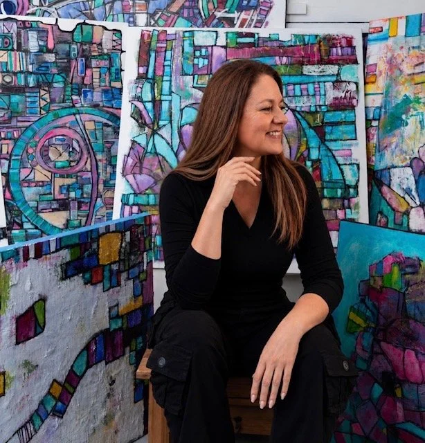 A woman with long brown hair, wearing a black shirt, smiling while sitting among colorful abstract paintings of cityscapes and geometric designs.
