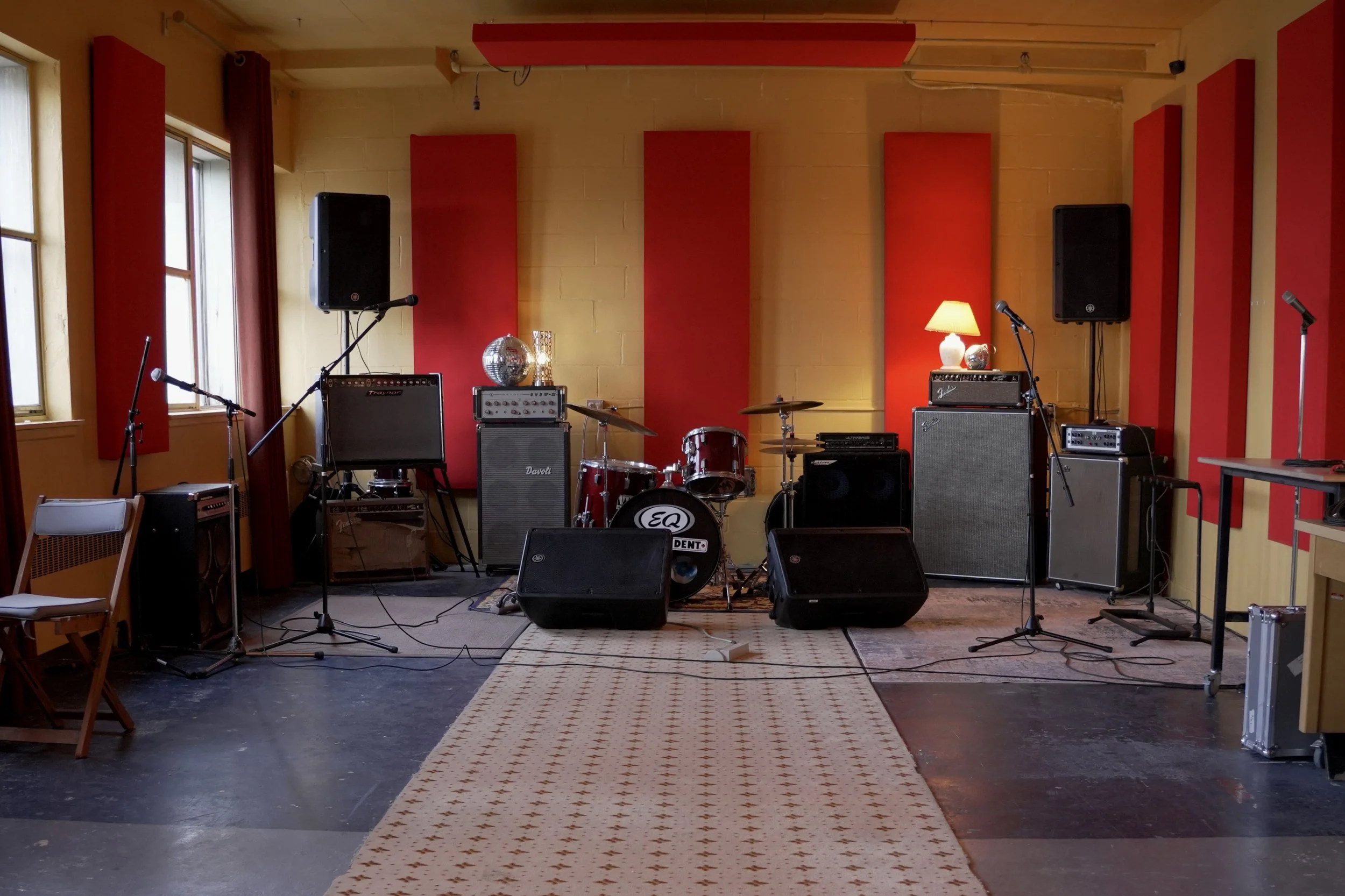 Scène de musique vide avec batteries, micros, haut-parleurs et amplificateurs dans une pièce aux murs jaunes et aux panneaux acoustiques rouges.