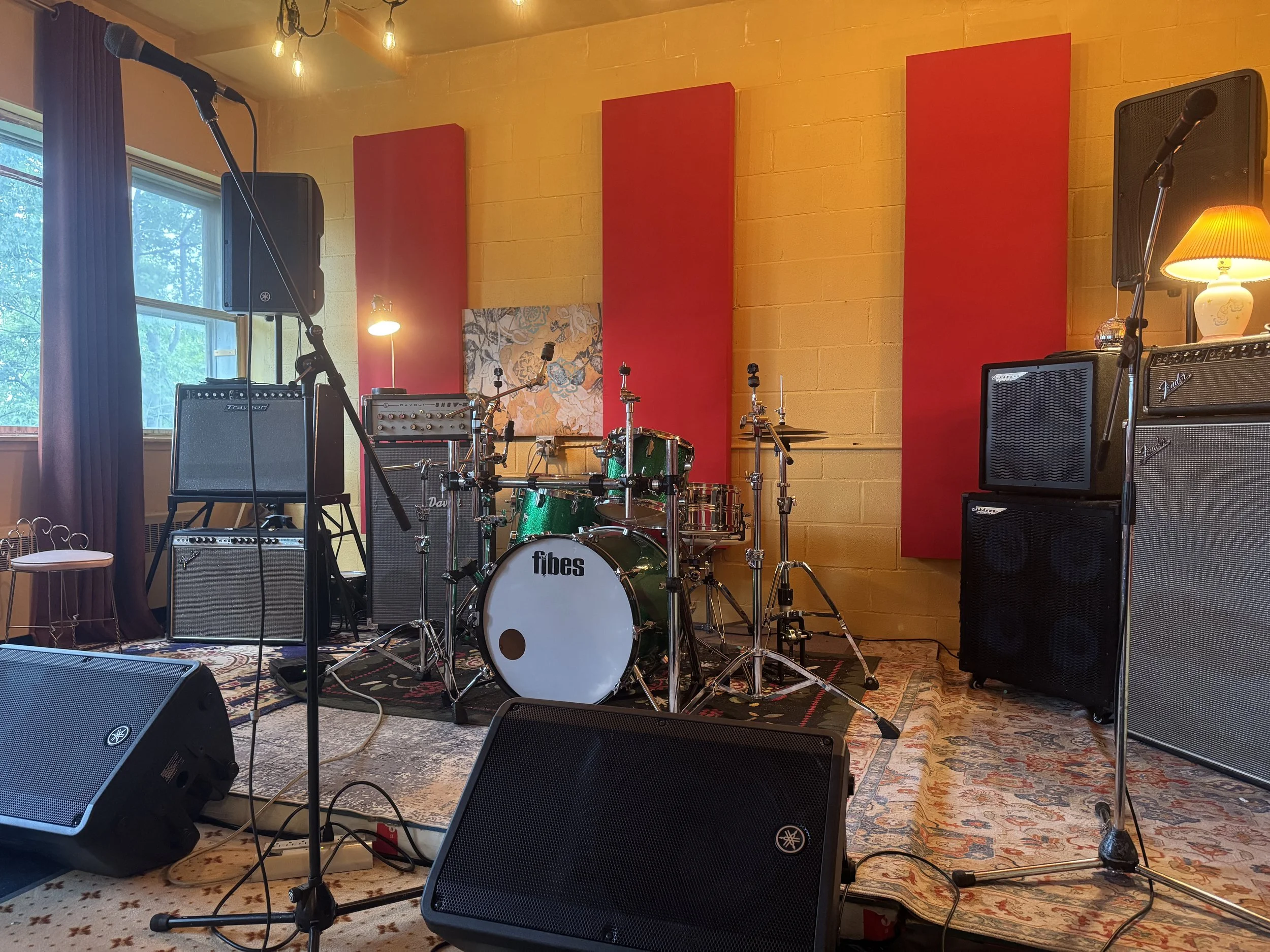 Scène musicale installée avec microphones, batterie, amplificateur et haut-parleurs dans une pièce aux murs jaunes, aux panneaux acoustiques rouges et aux fenêtres avec des rideaux rouges.