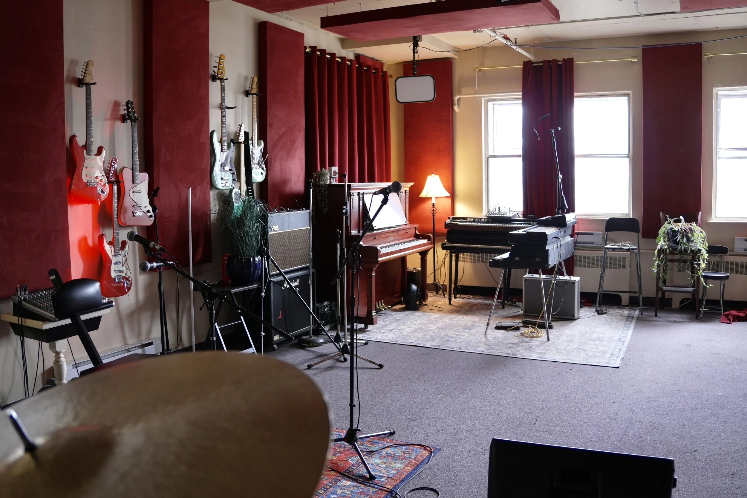 Une pièce avec des instruments de musique, dont un piano électrique Rhodes au premier plan, un clavier vintage en bois sur le dessus, et des guitares accrochées au mur en arrière-plan. Il y a des batteries, des amplificateurs et un mélangeur sur une table, avec une lampe sur le côté droit.