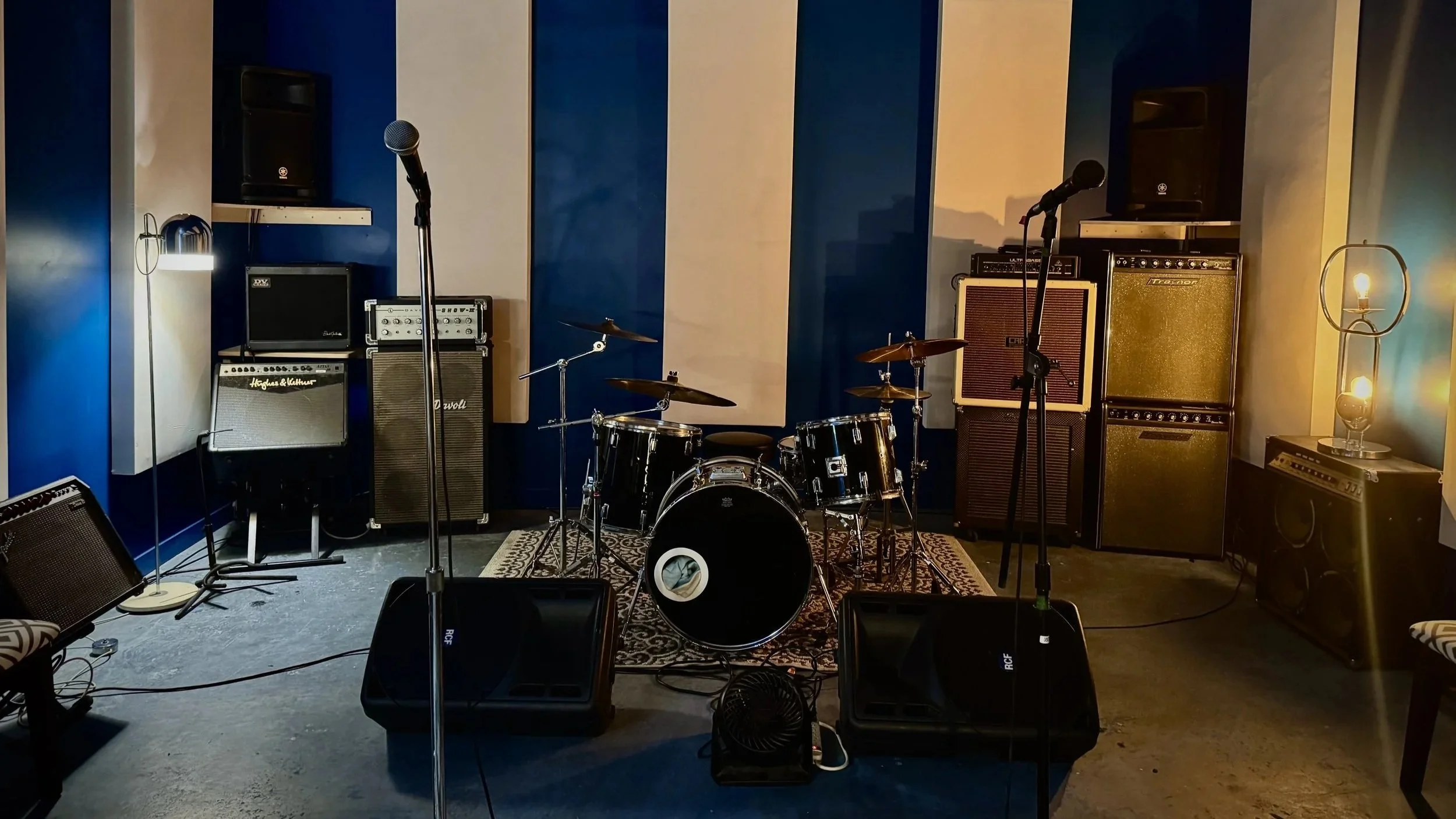 Une petite scène de musique avec batterie, guitares et micros, faiblement éclairée par un éclairage d’accent, installée dans un espace de spectacle chaleureux.