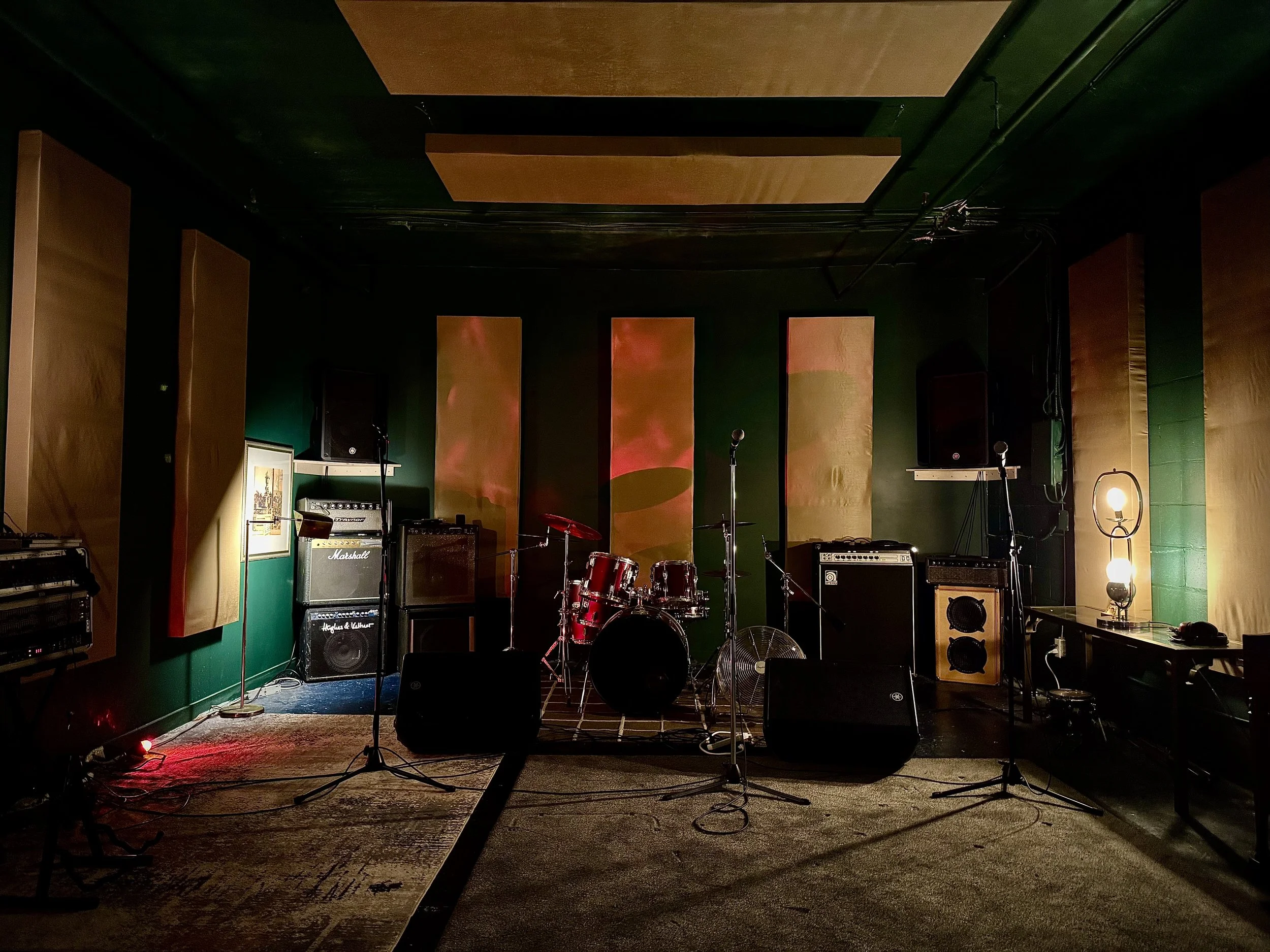 A dimly lit stage set up for a live music performance with musical instruments, amplifiers, mic stands, and a lamp, surrounded by soundproofing panels on the walls.