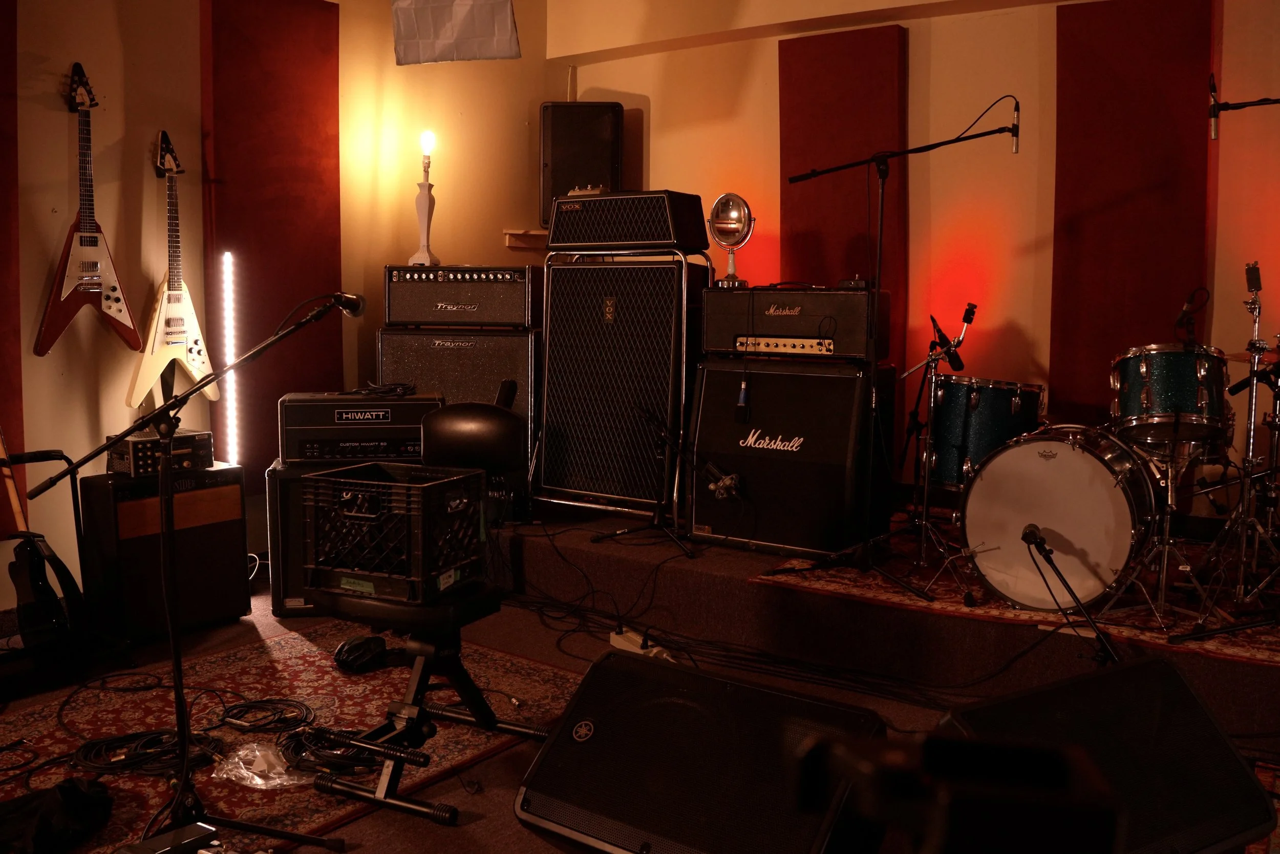 Music rehearsal room with guitars mounted on the wall, multiple amplifiers, microphones, drums, and other musical equipment, lit with warm lighting.