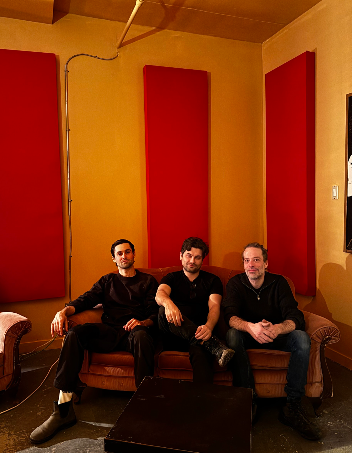 Trois hommes assis sur un divan vintage dans une pièce aux murs jaunes et aux panneaux acoustiques rouges.