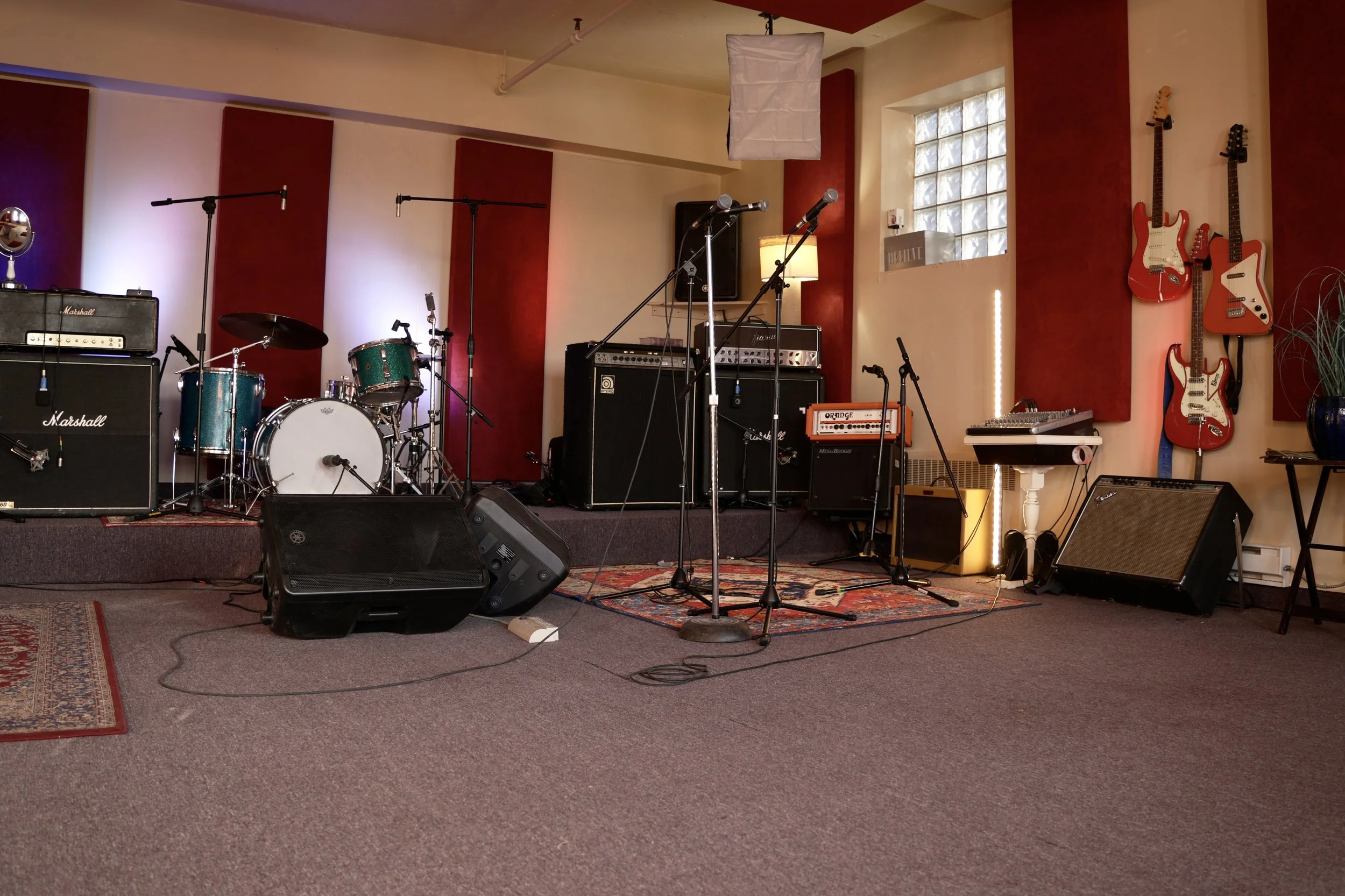 Un studio de répétition musicale avec des guitares suspendues sur des panneaux muraux rouges, de l’équipement audio incluant des amplificateurs Marshall et Fender, une batterie, des pieds de micro et des lumières aux teintes violettes et roses.