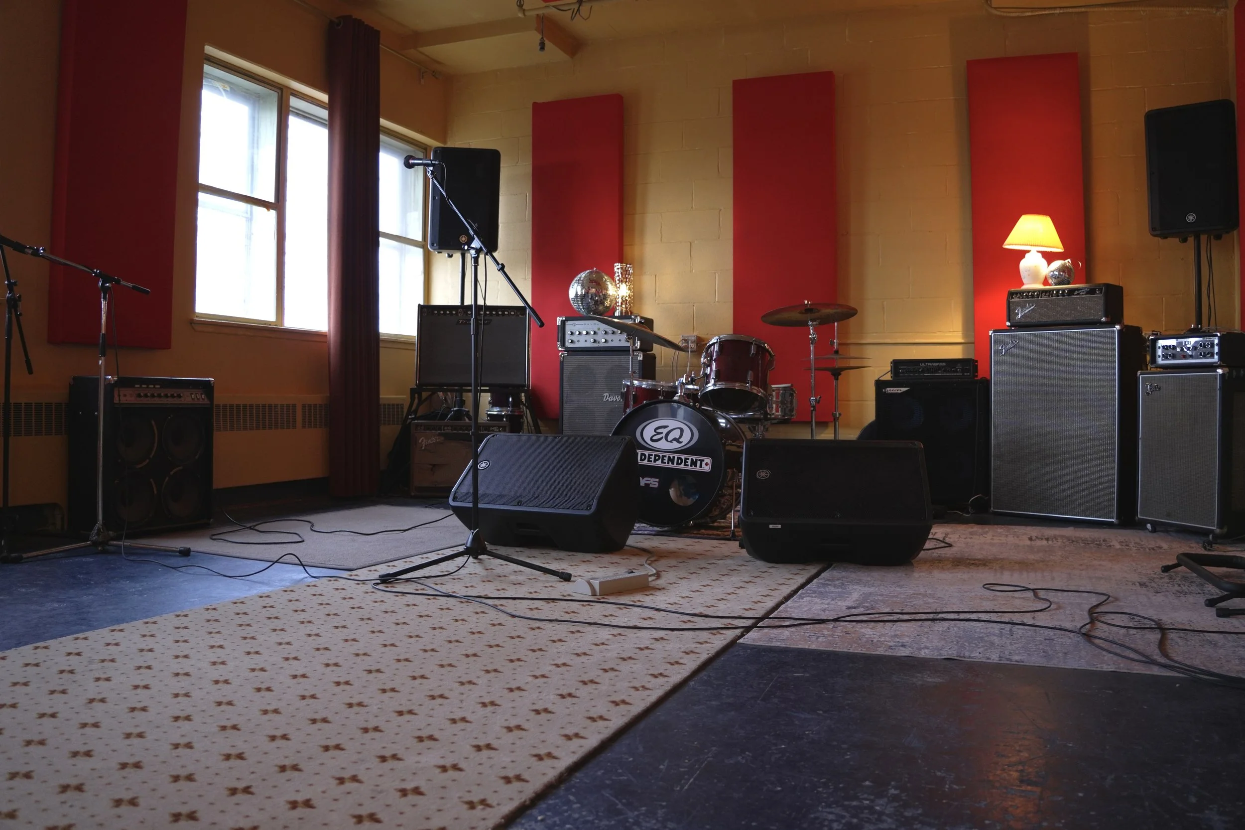 Scène musicale installée avec microphones, batterie, amplificateur et haut-parleurs dans une pièce aux murs jaunes, aux panneaux acoustiques rouges et aux fenêtres avec des rideaux rouges.
