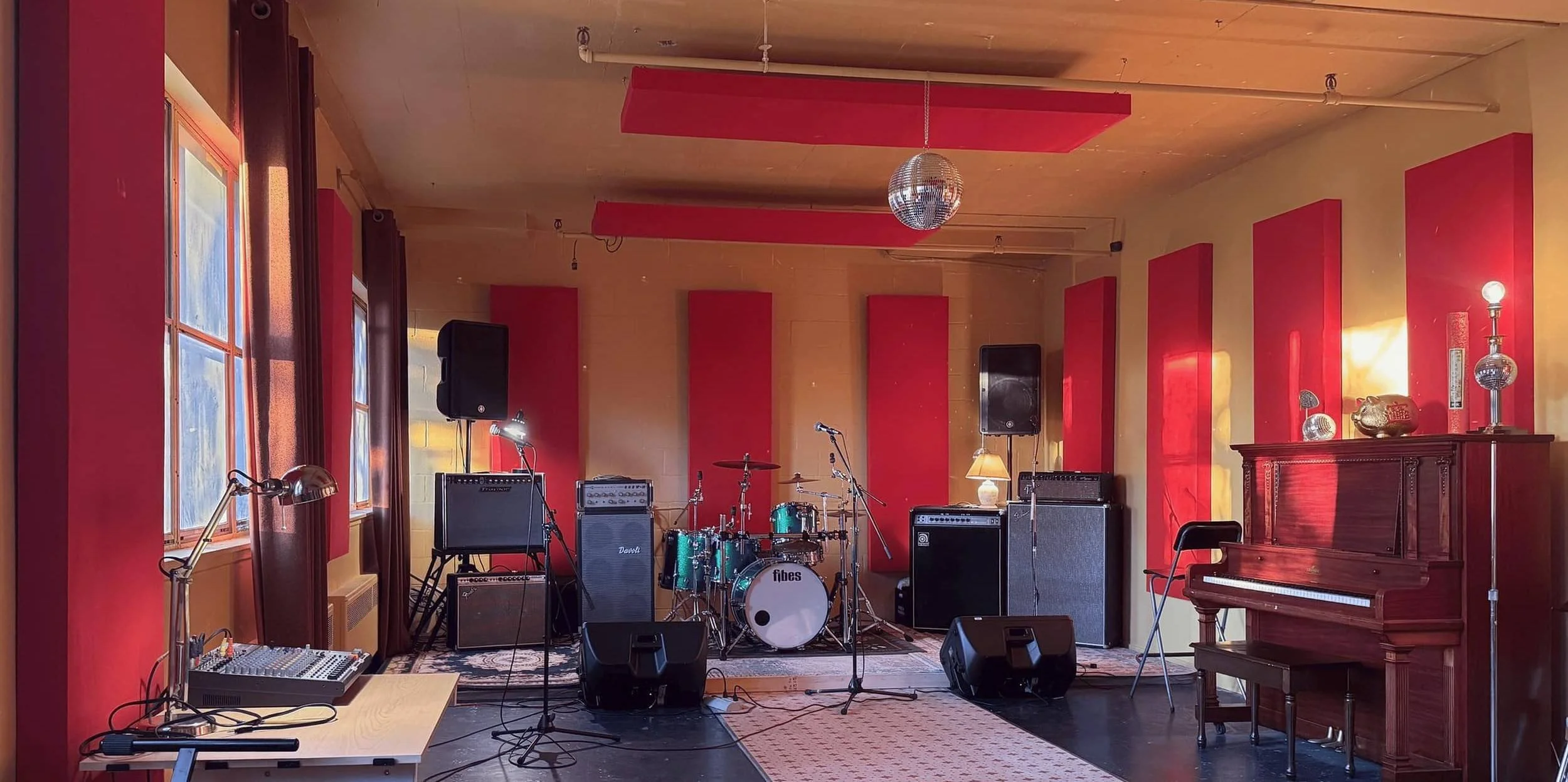 Salle de répétition lumineuse au studio NOW de Montréal, avec batterie, amplis, panneaux acoustiques rouges et grandes fenêtres.