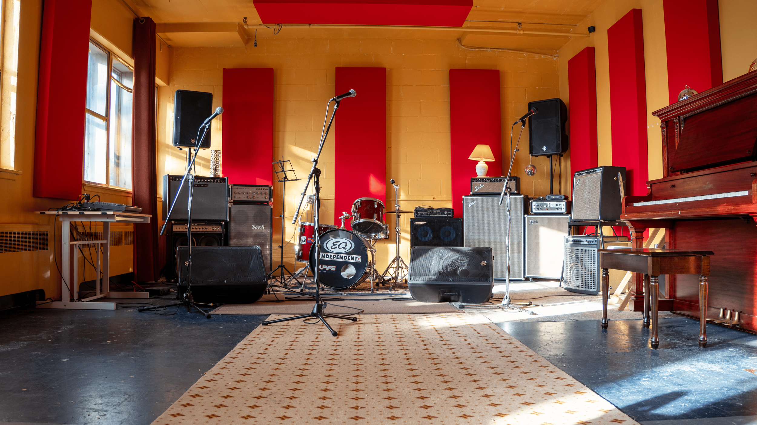 Music stage set up with microphones, drum kit, amplifier, and speakers in a room with yellow walls, red acoustic panels, and windows with red curtains.