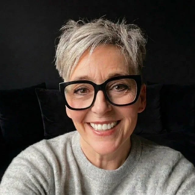 A smiling woman with short gray hair wearing black glasses and a light gray sweater, sitting against a black background.