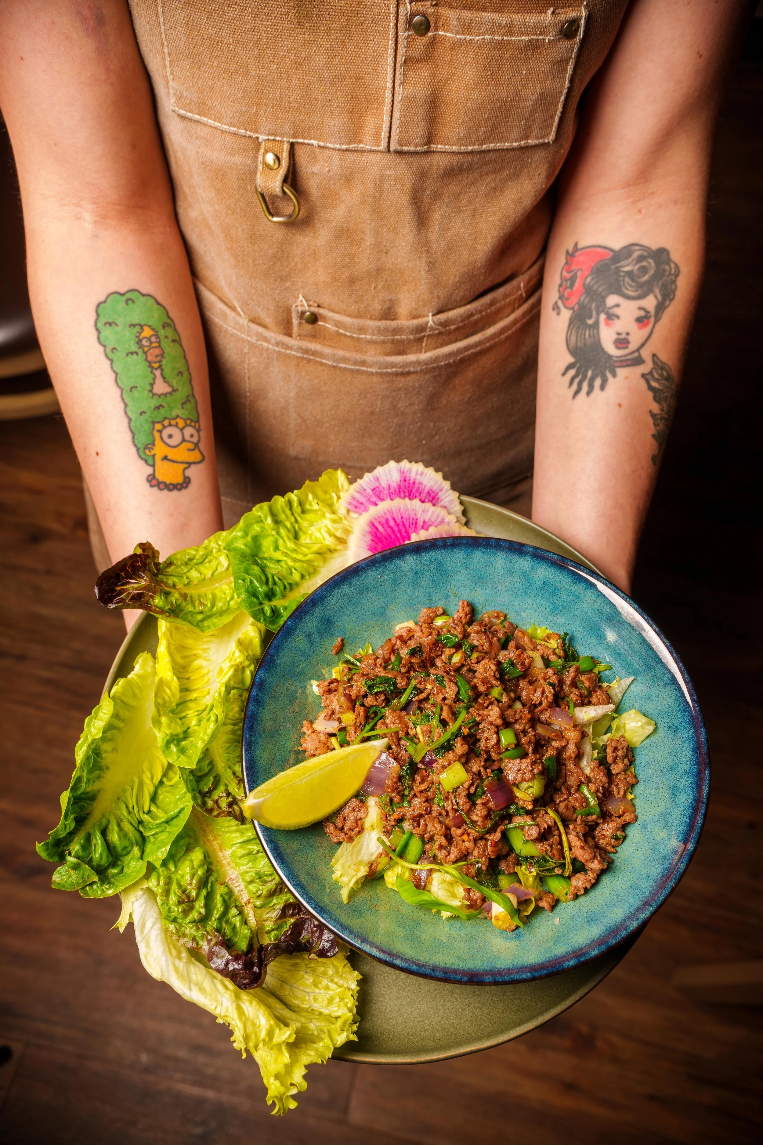 A person wearing a brown apron with tattoos on both forearms, one of Homer Simpson and the other of a woman with dark hair and a rose in her hair, is holding a bowl of stir-fried ground meat with green onions and a lemon wedge on a bed of lettuce and radish slices.