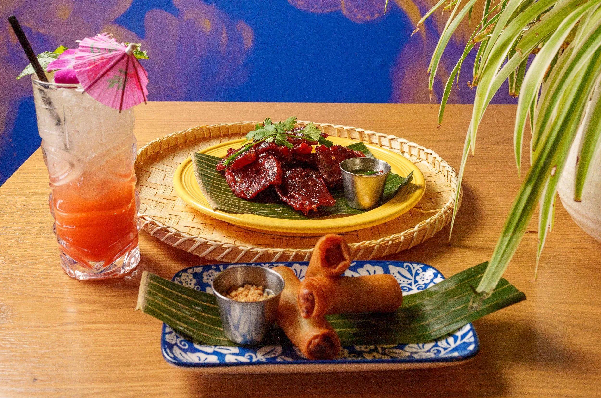 A tropical cocktail with a pink paper umbrella and two straws next to a plate of Mexican food, including crispy spring roll appetizers, spicy beef with cilantro, and a small container of dipping sauce, all on a bamboo tray on a wooden table.