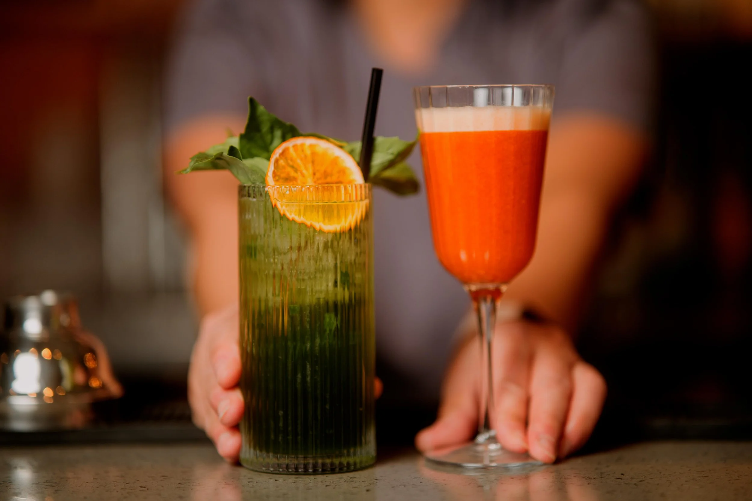 A person holding a green cocktail garnished with an orange slice and mint leaves, and another person holding a tall glass of orange-colored drink with a straw.