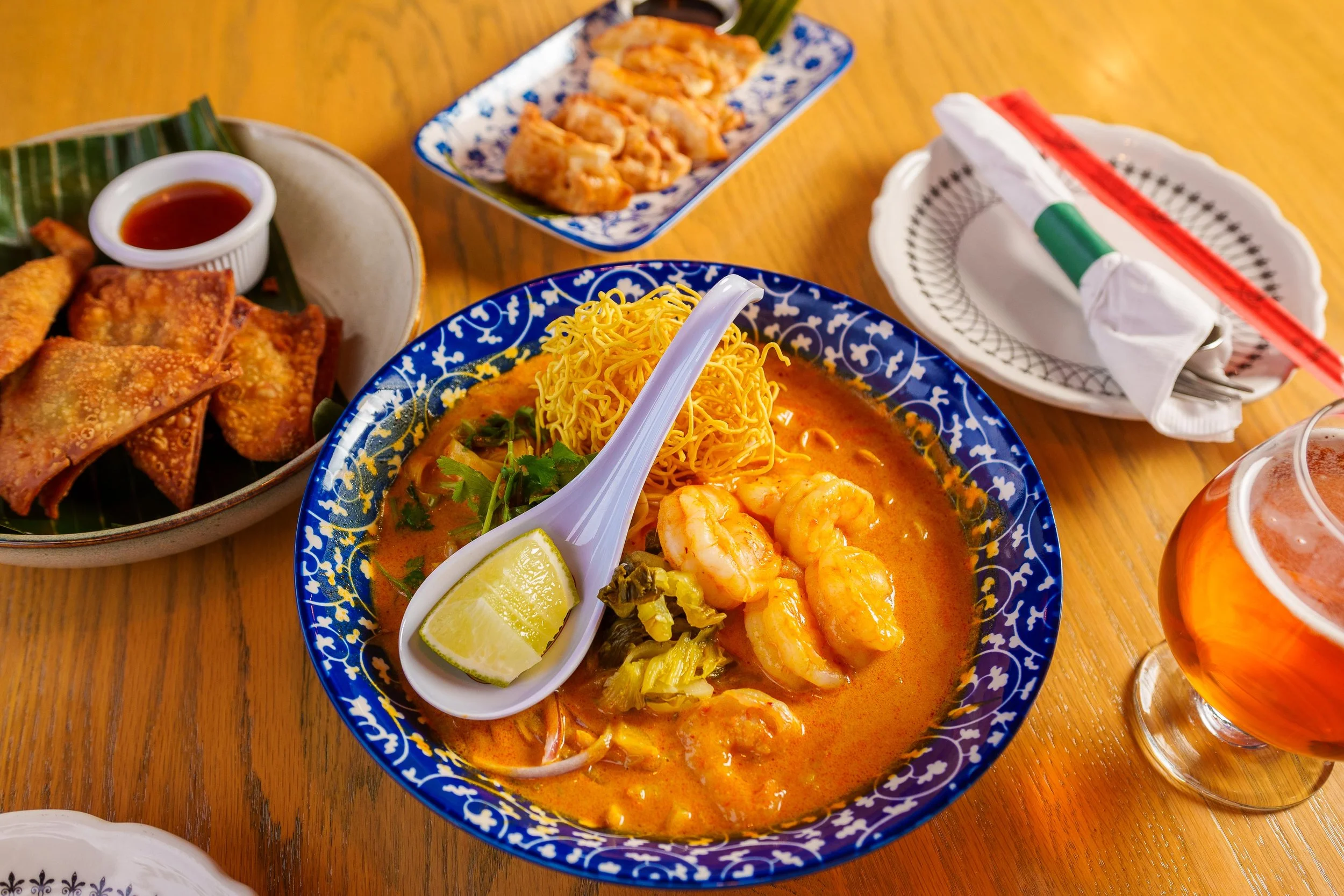 A close-up of Thai dishes including shrimp in curry with lime wedges, crispy spring rolls with dipping sauce, and a plate of satay skewers on a wooden table.