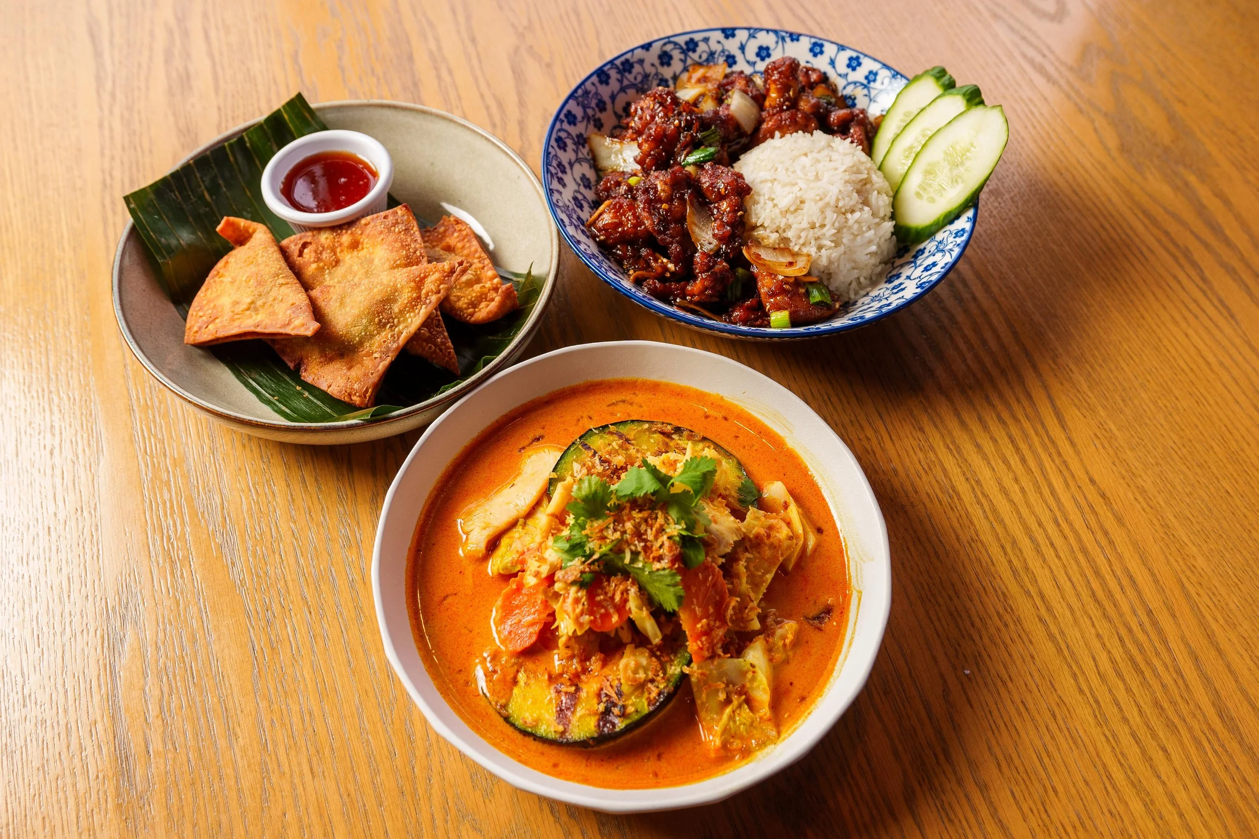 Three bowls of Asian cuisine on a wooden table. The first bowl contains spring rolls with dipping sauce, the second bowl contains beef and rice with cucumber slices, and the third bowl contains a spicy vegetable curry with cilantro.