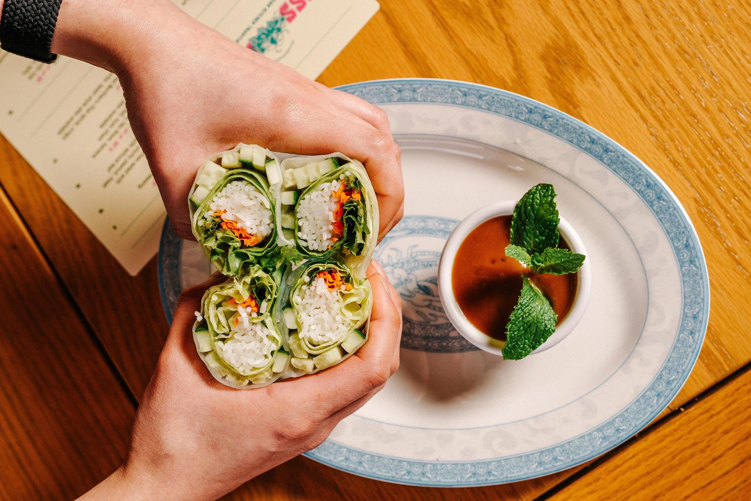 Someone holds a fresh spring roll filled with rice noodles, vegetables, and herbs. A bowl of dipping sauce garnished with mint leaves is on a blue and white plate on a wooden table, with a menu partially visible in the background.