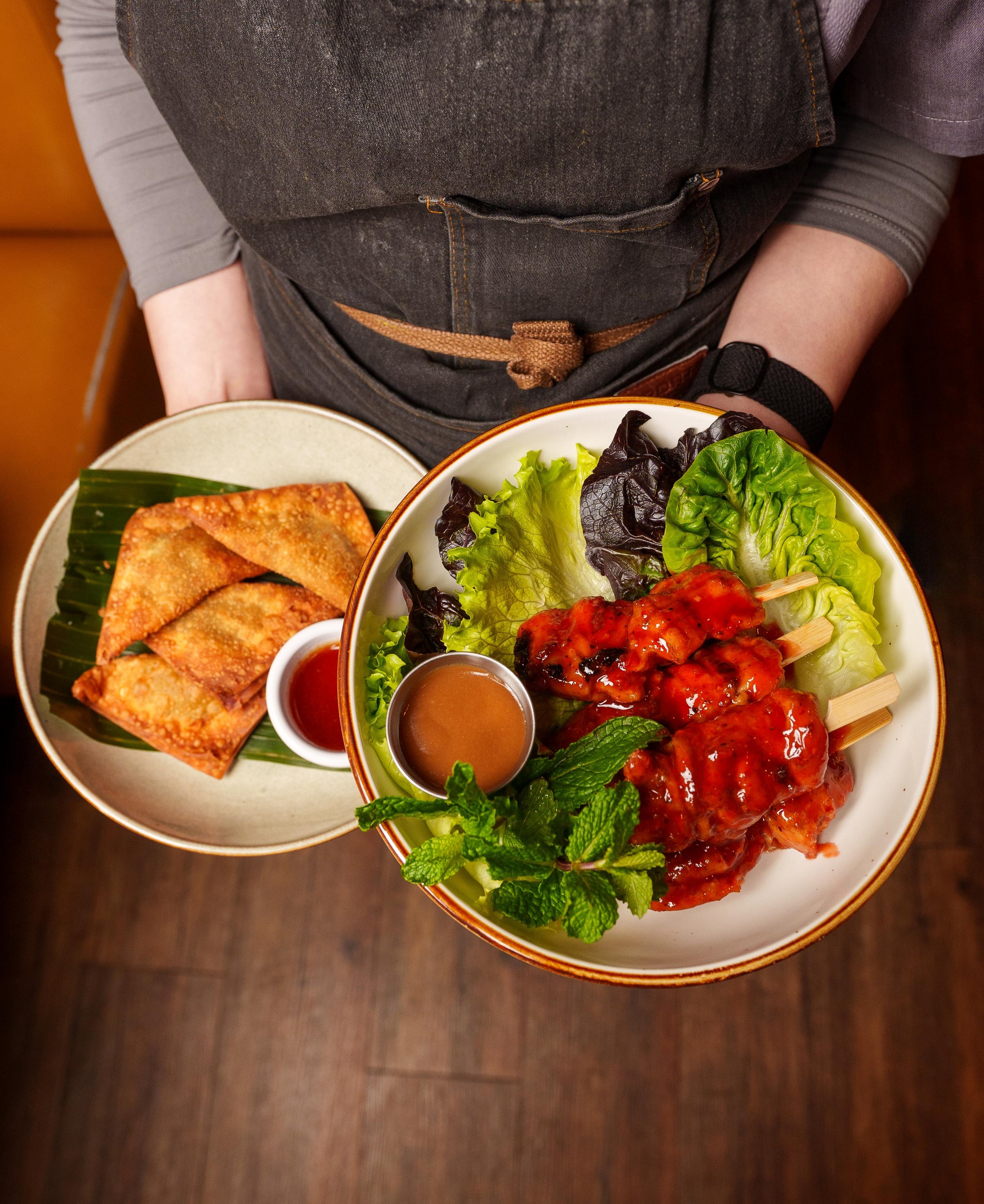 Top view of a bowl filled with leafy greens, grilled chicken skewers, and mint leaves. Next to the bowl, there is a plate with fried spring rolls and two small containers of dipping sauces, all on a dark wooden table.