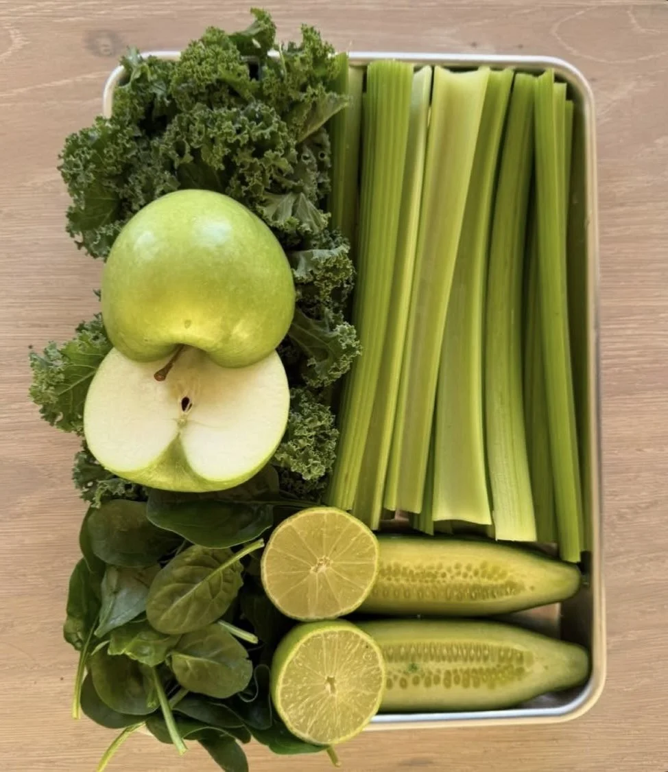 A tray of fresh green vegetables and fruits including kale, celery sticks, a whole green apple, apple halves, spinach, a halved lemon, and cucumber halves on a wooden surface.